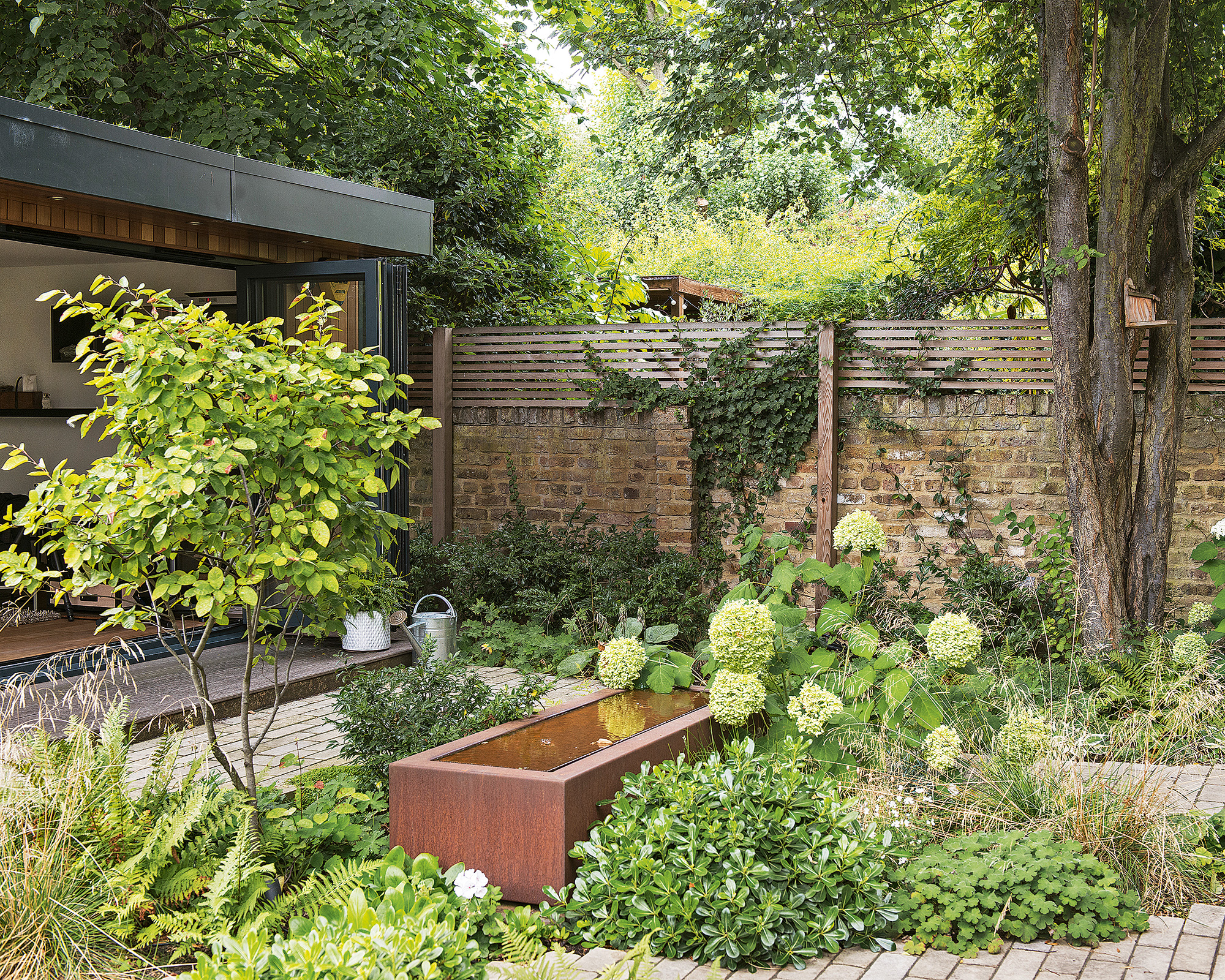 Petit jardin avec mur de briques, plancher en briques, récipient d'eau en métal, fleurs, buissons, arbres