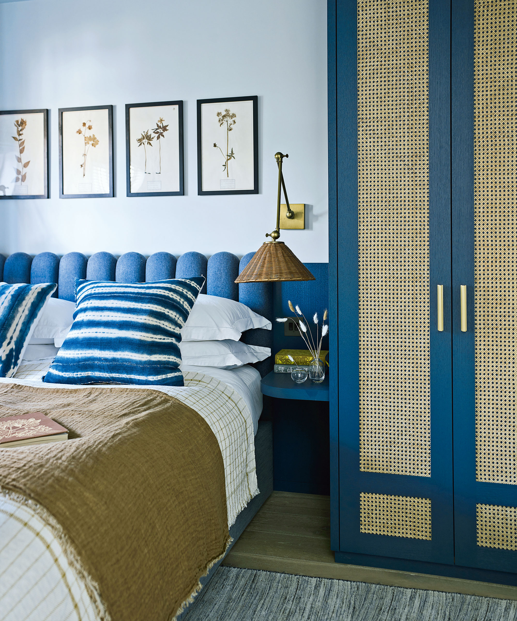 Chambre peinte en bleu pâle, tête de lit festonnée indigo, armoire avec portes en rotin dans la teinte indigo assortie, linge de lit blanc avec coussins rayés blanc et indigo, jeté marron, œuvre d'art assortie au-dessus du lit montrant des fleurs pressées, petite étagère de chevet, applique murale avec abat-jour en osier tressé au-dessus de.
