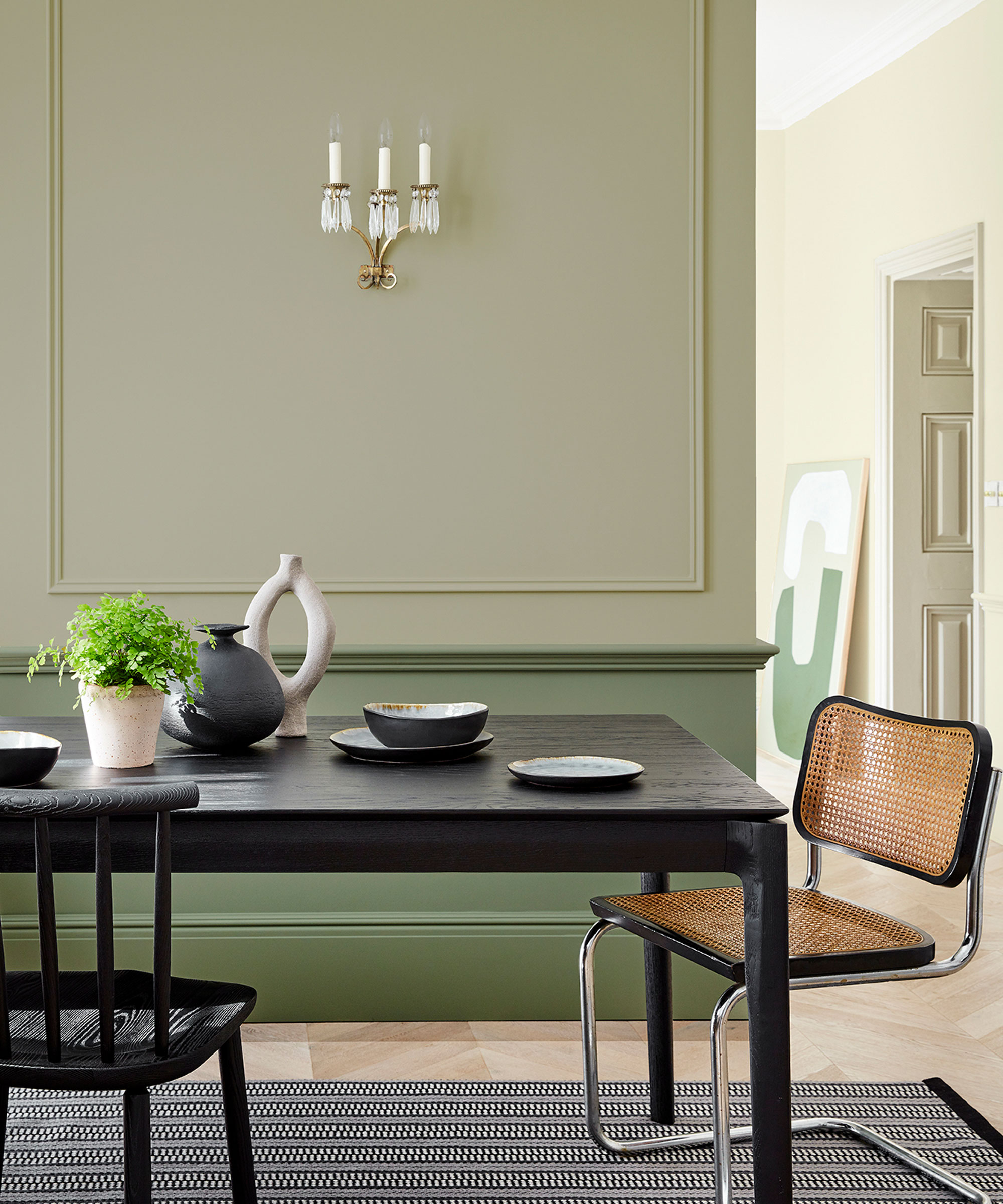 Salle à manger verte peinte en deux tons de petit vert, lambris muraux sur les murs, table à manger en bois noir, chaise cesca en noir et rotin, table à manger habillée d'ornements, d'assiettes et de plantes, candélabres au mur