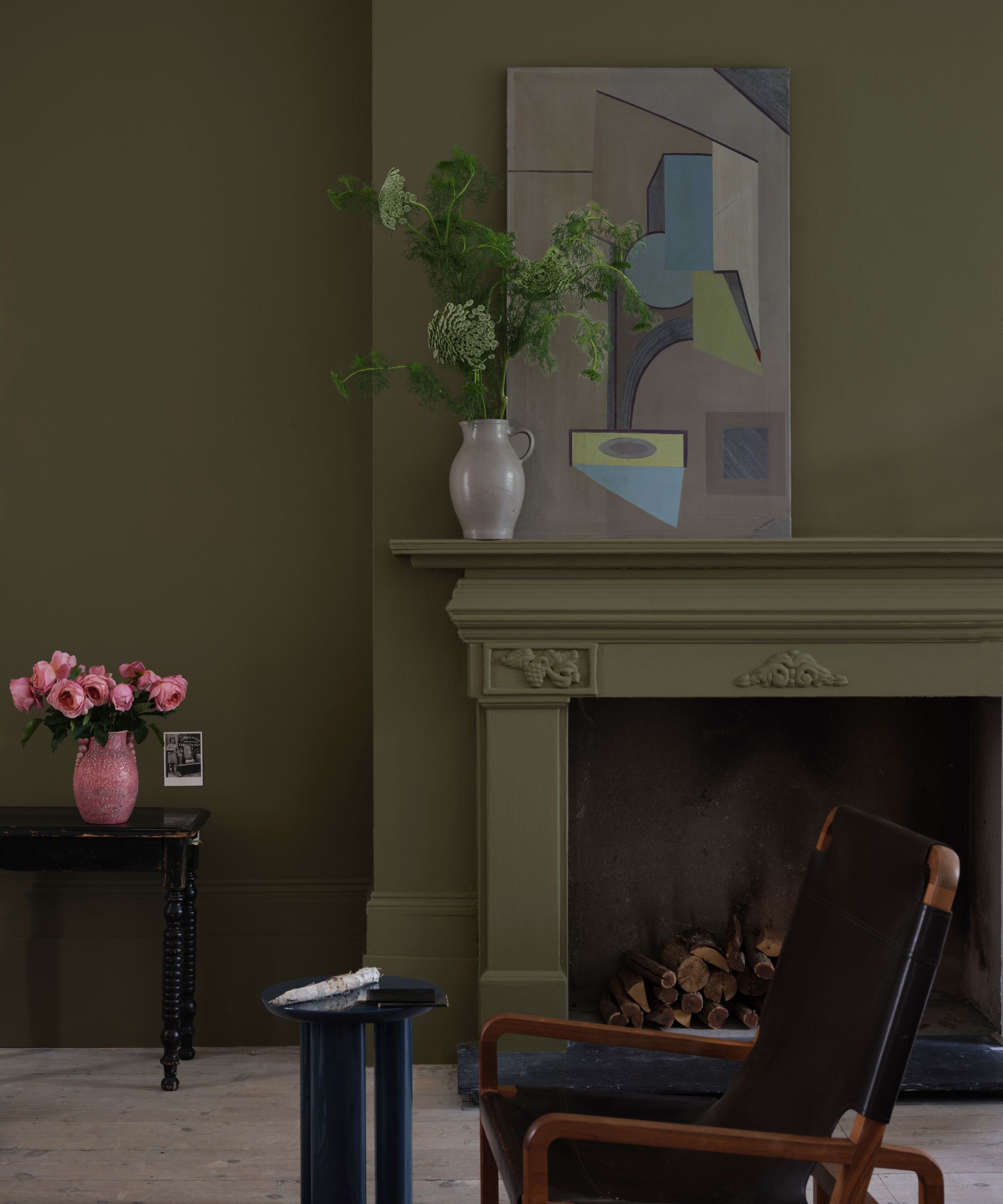 salon sombre avec murs verts olive, cheminée et vase de feuillage et illustrations sur le manteau, fauteuil noir au premier plan