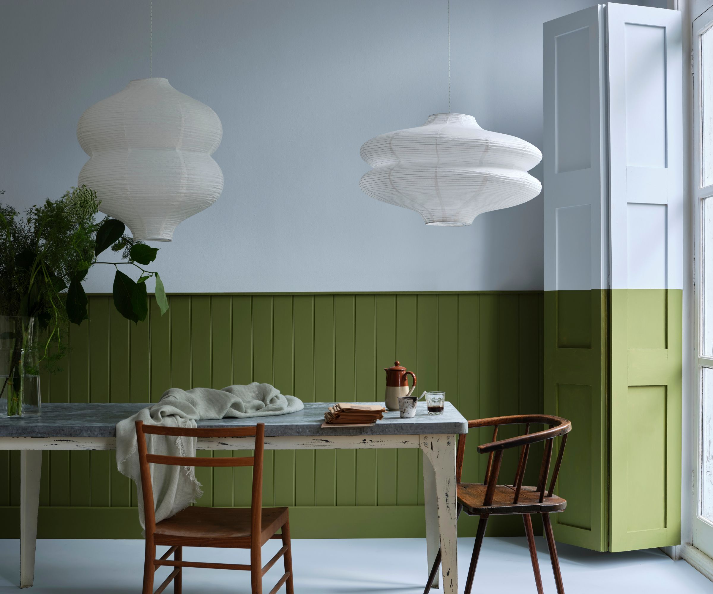 Espace à manger avec une moitié supérieure bleu clair des murs et des panneaux verts foncés sur le bas. table à manger avec deux chaises en bois vintage et deux feux de suspension en papier blanc