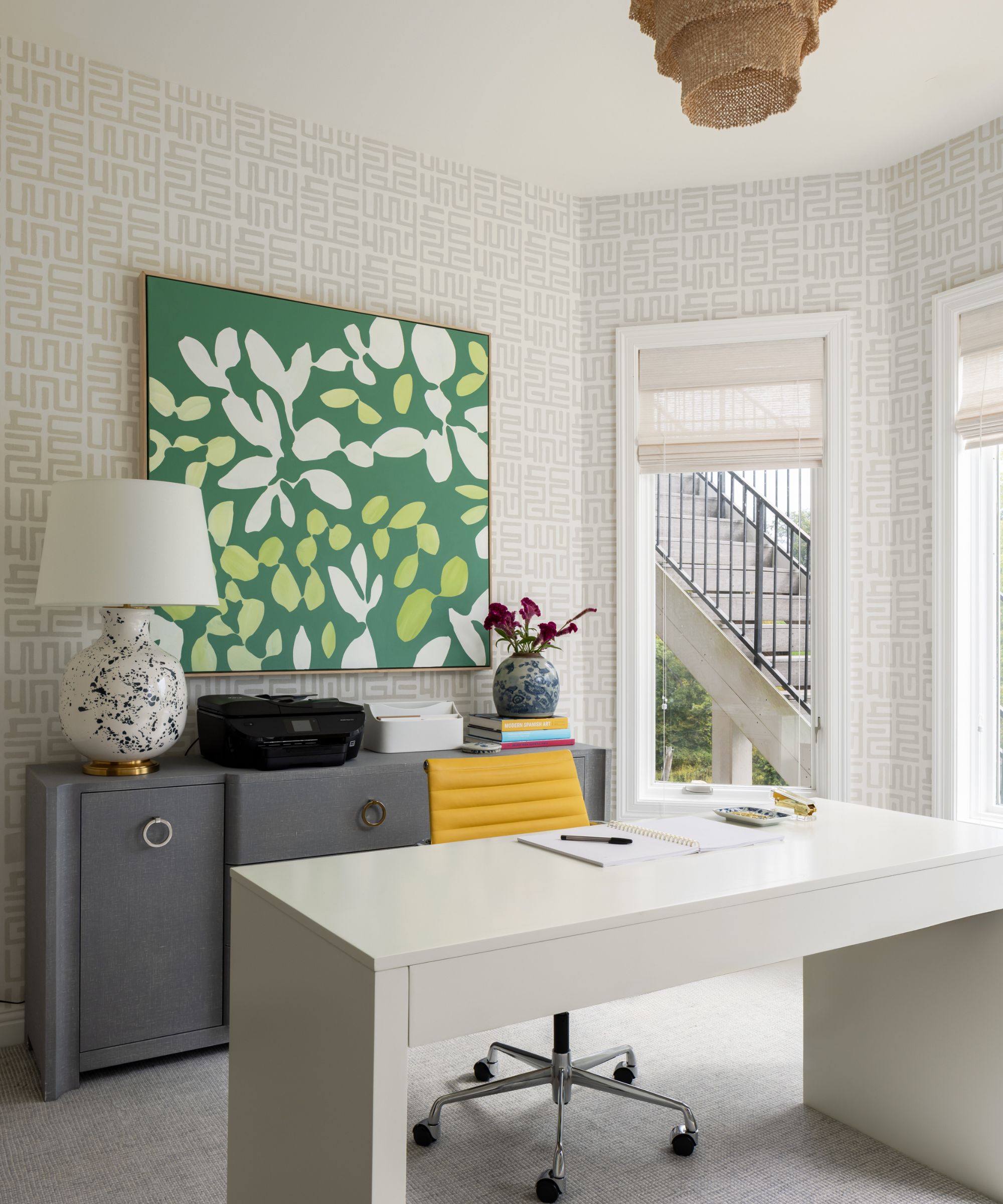 Bureau à domicile avec papier peint à motifs neutres, garniture blanche et bureau blanc avec chaise pivotante jaune, œuvres d'art floral vertes sur mur et armoire grise