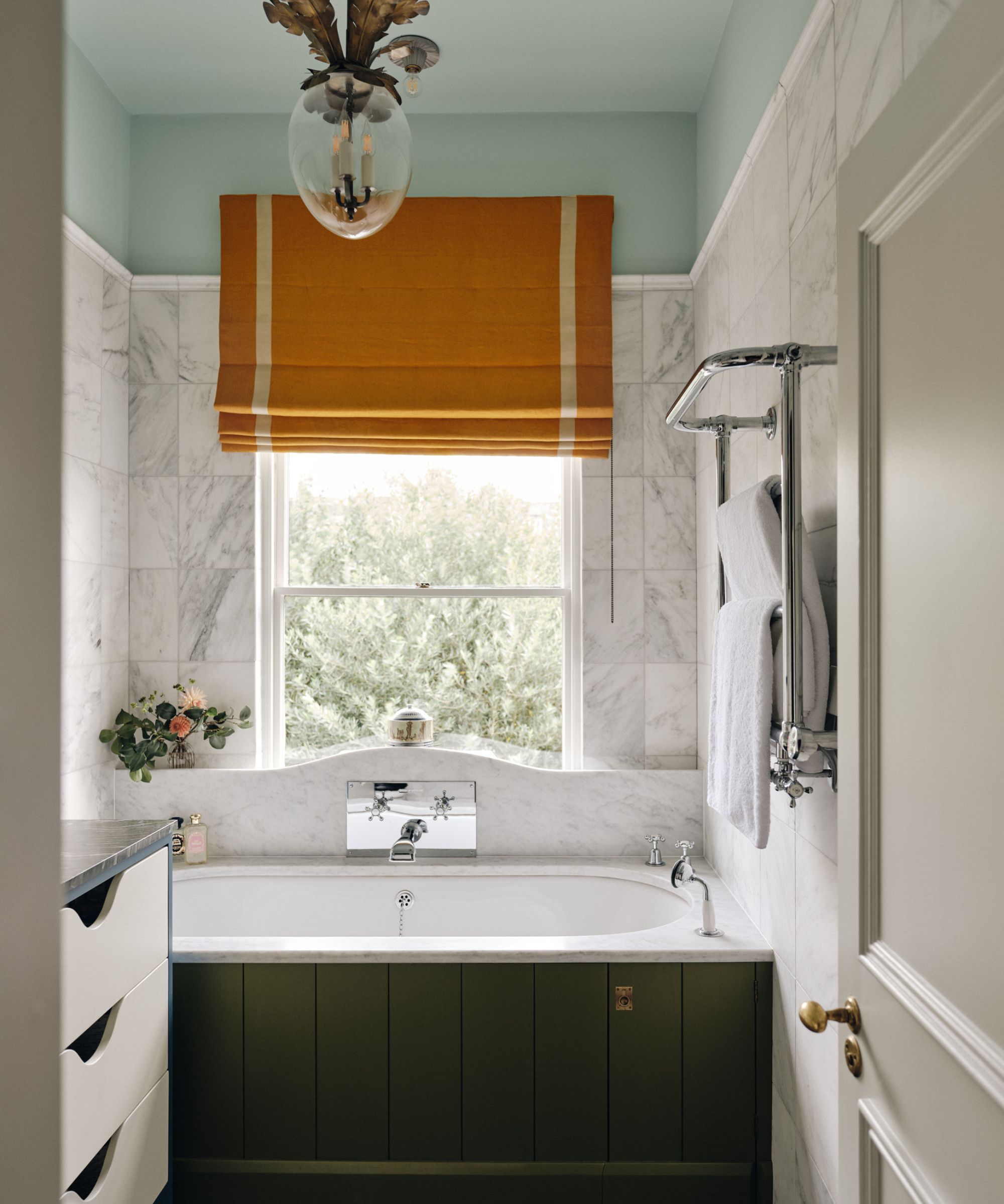 Salle de bain neutre avec des murs en marbre carrelé et une baignoire en lambris verte, un plafond bleu pâle et une fenêtre orange aveugle