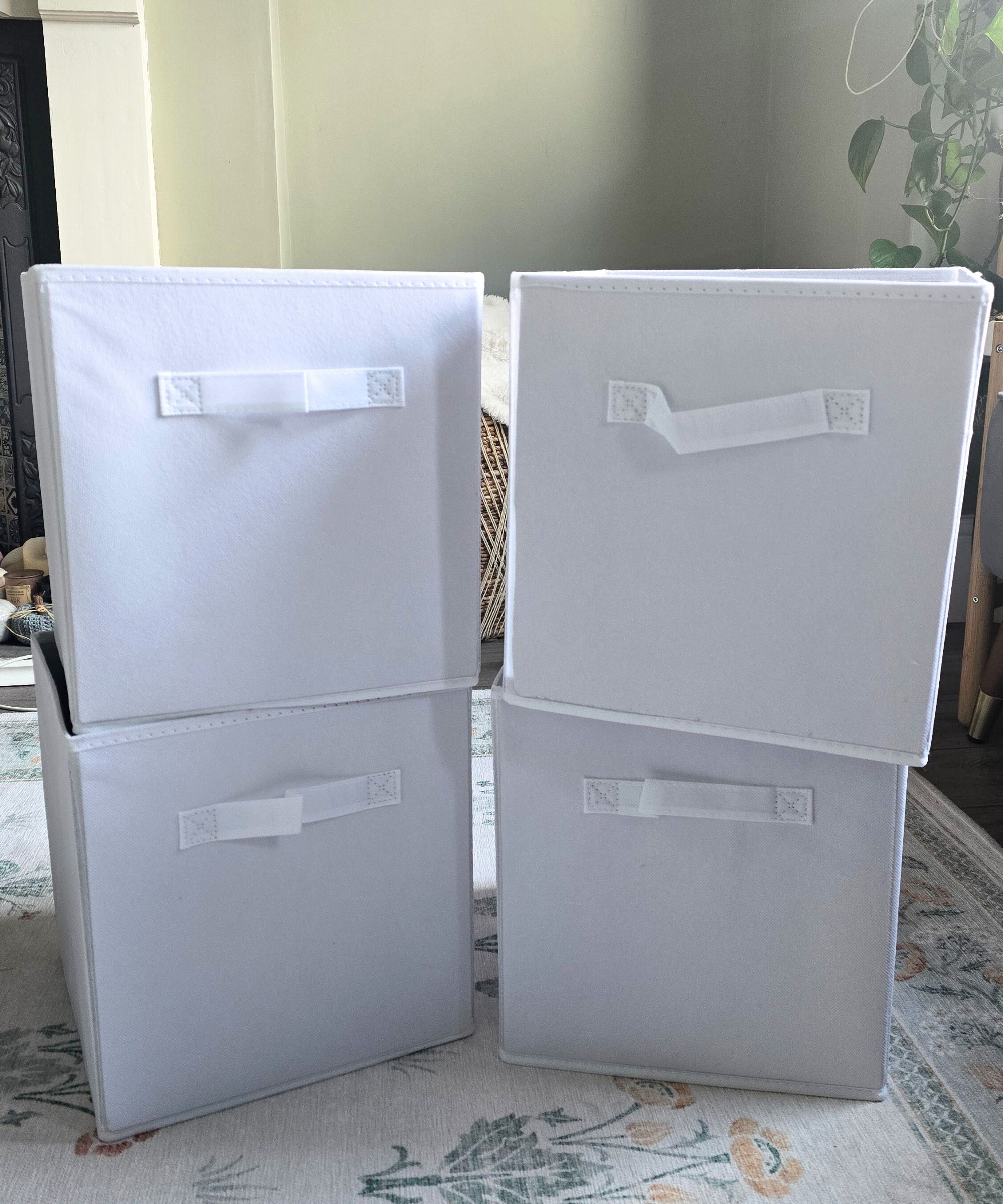 Quatre stocks de cubes de tissu blanc empilés en deux tours, avec un mur vert et une plante de lierre du diable derrière eux et un tapis floral vert et blanc sous eux