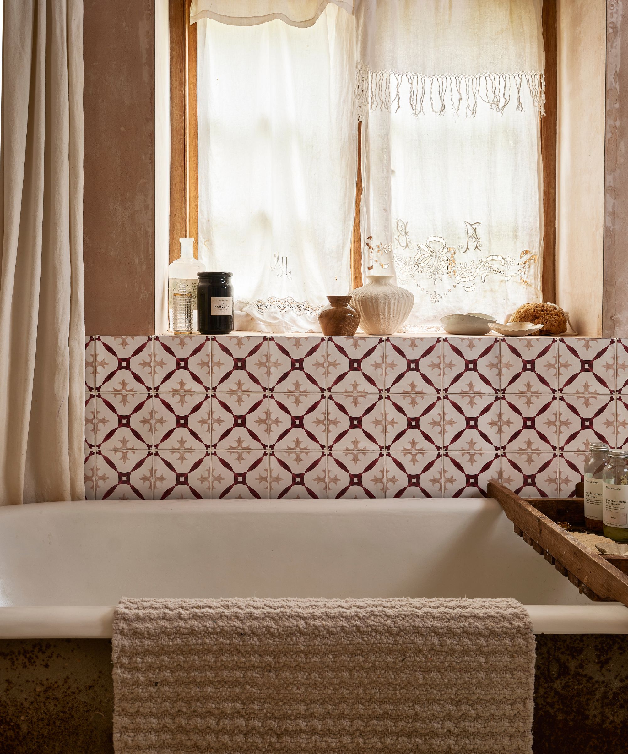 Une salle de bain avec des murs lavés marron, une fenêtre avec des rideaux blancs, des carreaux à motifs brun blanc et foncé, et une baignoire avec un plateau de bain en bois et une serviette beige suspendue au bord de celui-ci