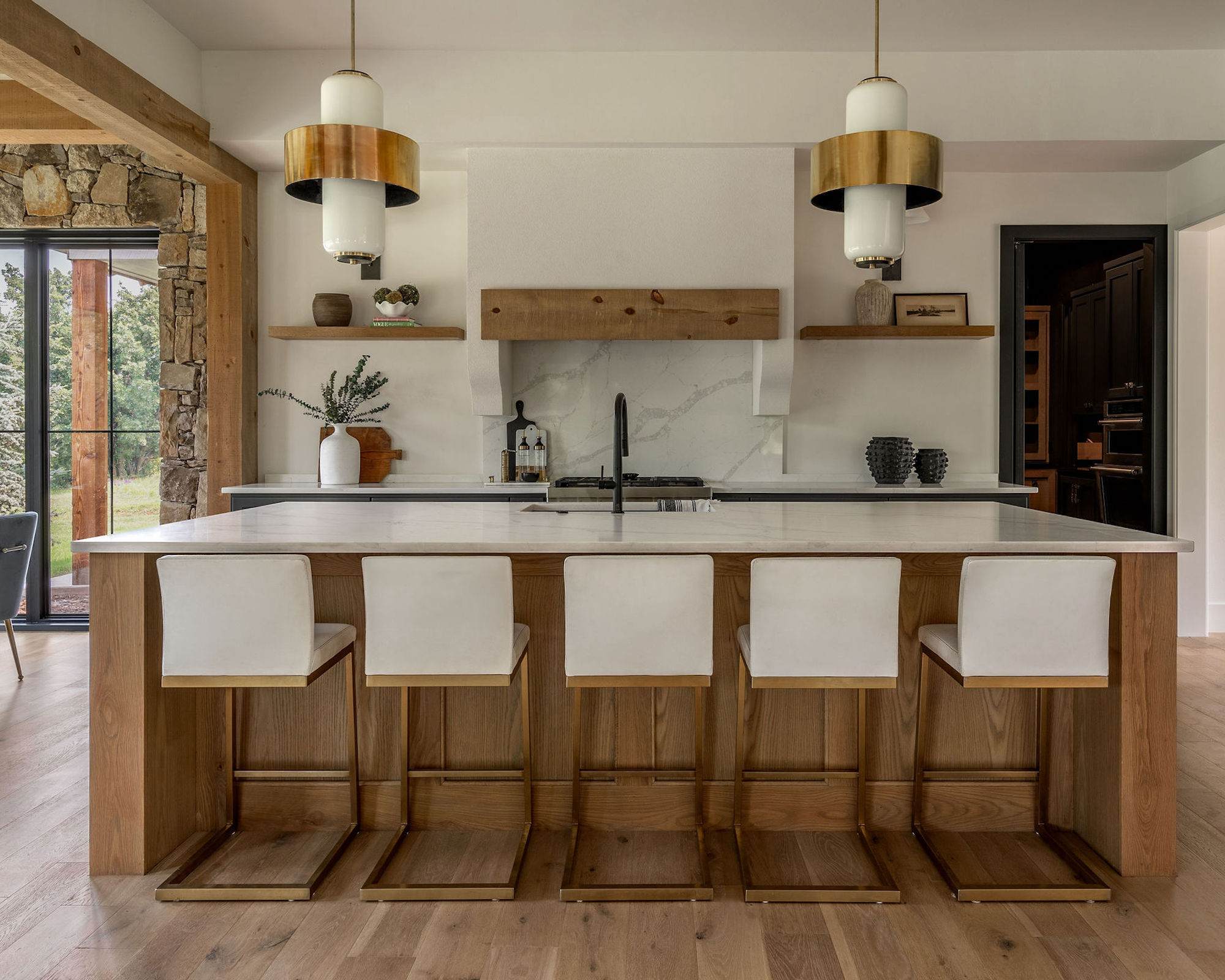 Cuisine avec îlot en bois et chaises de bar, comptoirs veinés, gamme avec dosseret, murs blancs et plancher en bois