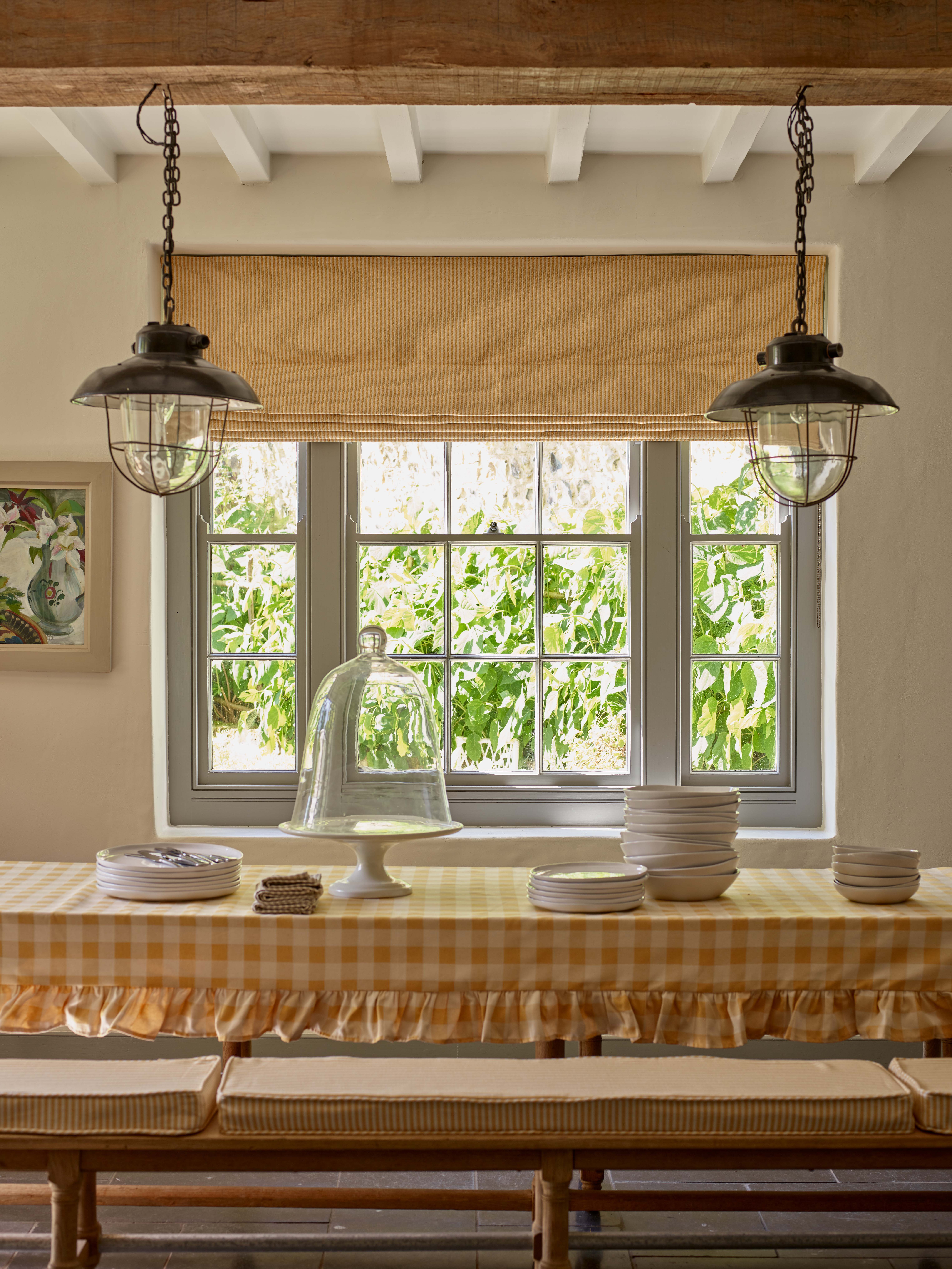 Idée de décoration de printemps de la ferme avec nappe de table en vigueur jaune avec fioritures, places de banc à rayures jaunes et aveugles assortis, pendentif de style pêcheur, poutres, œuvres d'art, assiettes et supports à gâteau sur table