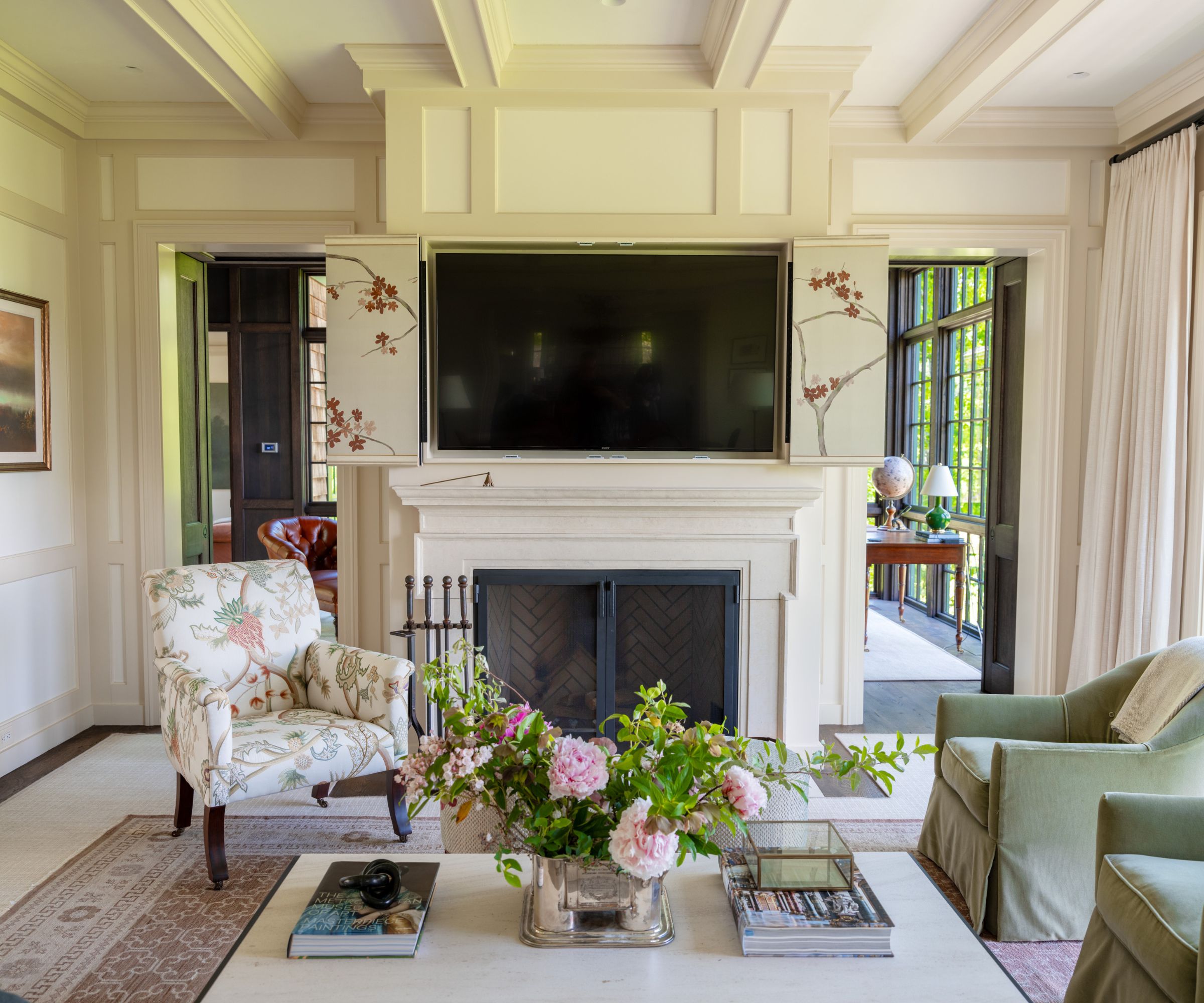 salon traditionnel avec murs en lambris crème, cheminée, fauteuils verts et fauteuil à motifs, grande table basse avec des fleurs roses en vase