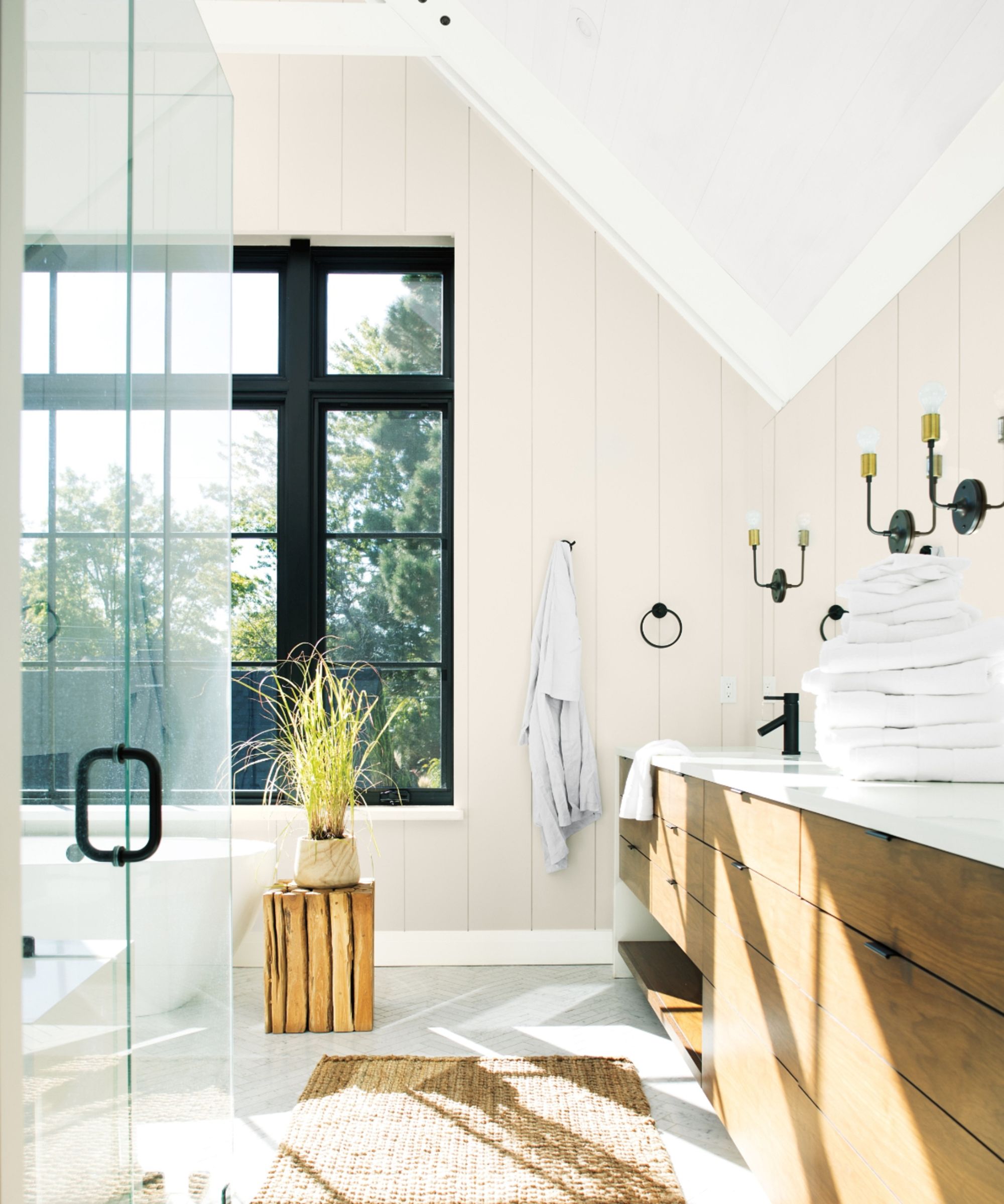 Salle de bain avec murs en lambris neutres, plafond en pente blanc, cadre de fenêtre noir, vanité en bois et coin de la douche en verre