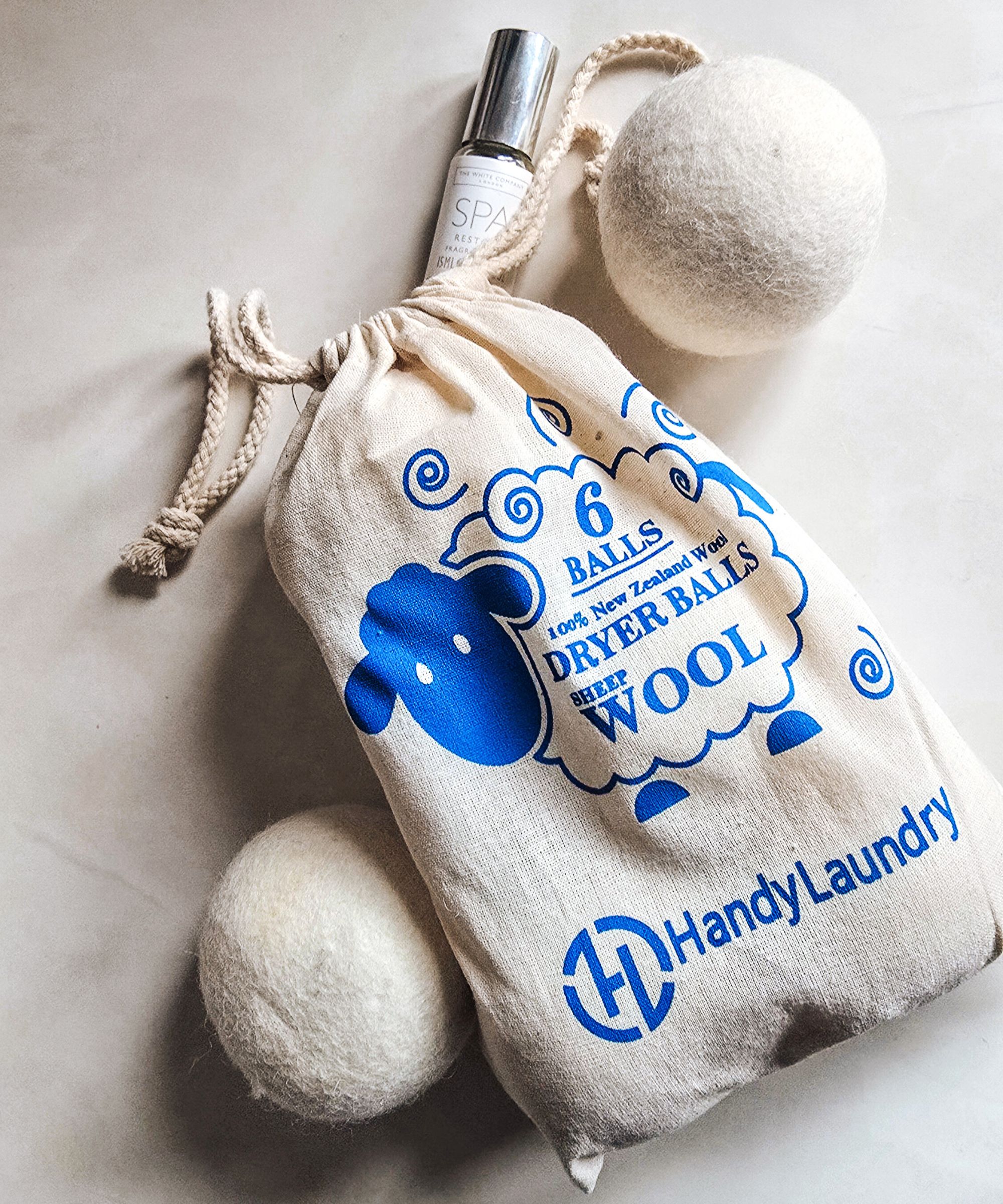 Un sac beige avec une illustration de mouton bleu dessus, avec deux boules de séchage en laine beige autour de lui et une bouteille d'huile essentielle en haut, allongé sur une surface blanche