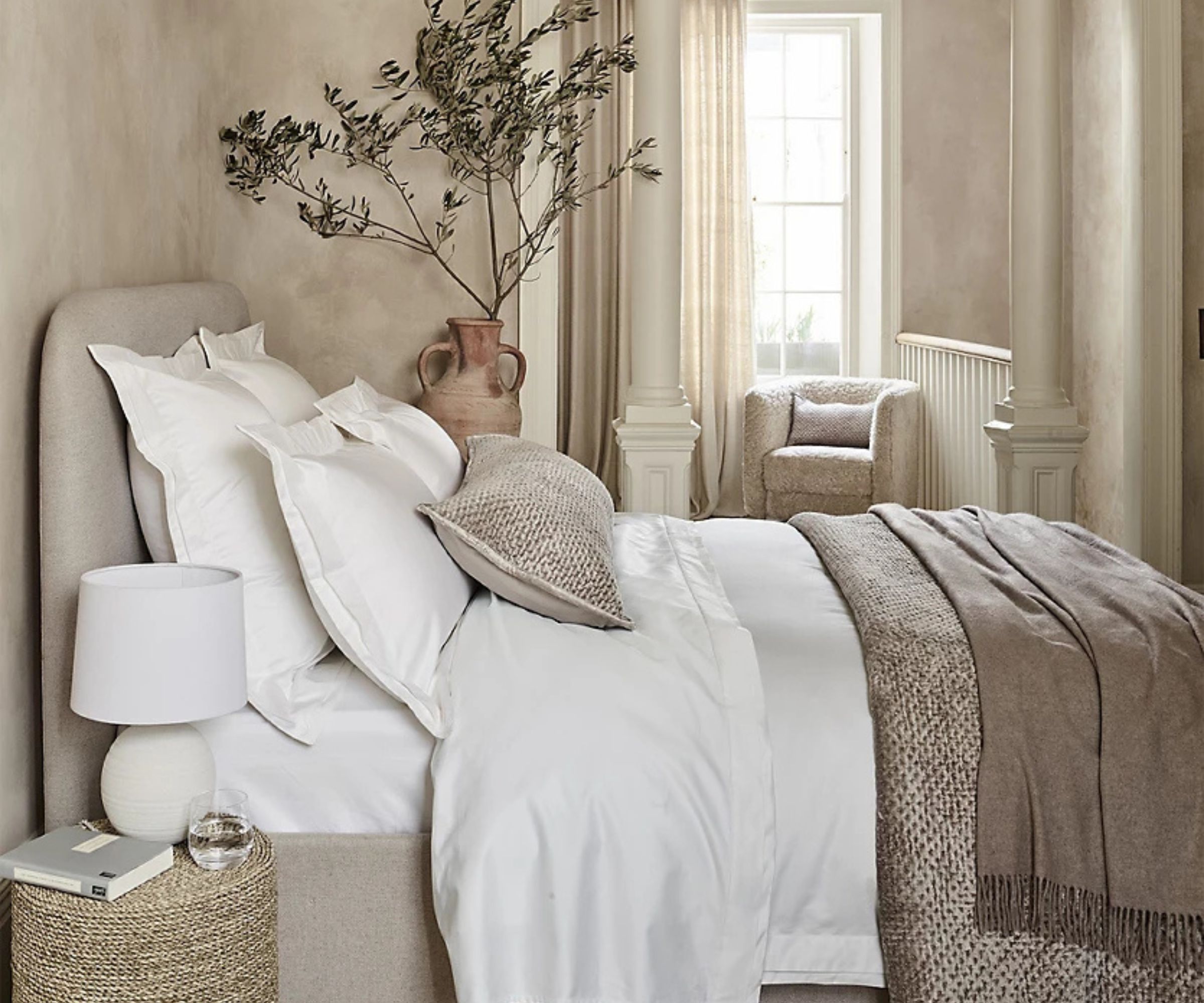 Collection de linge de lit Kingham sur un lit contre un mur beige avec une plante d'intérieur en arrière-plan.