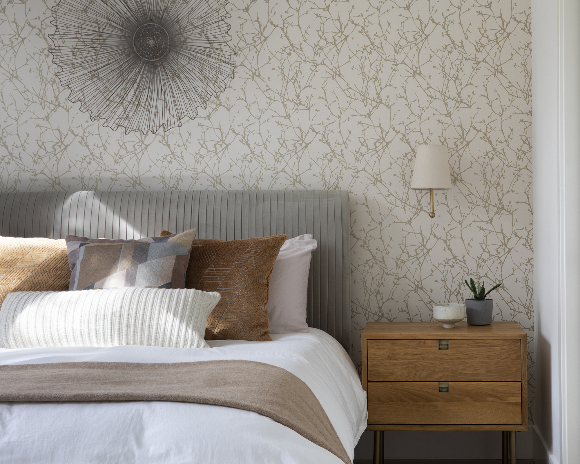 Chambre avec lit habillé de lin blanc et jet neutre et oreillers, table de chevet, lumière murale, papier peint à motifs et décoration murale