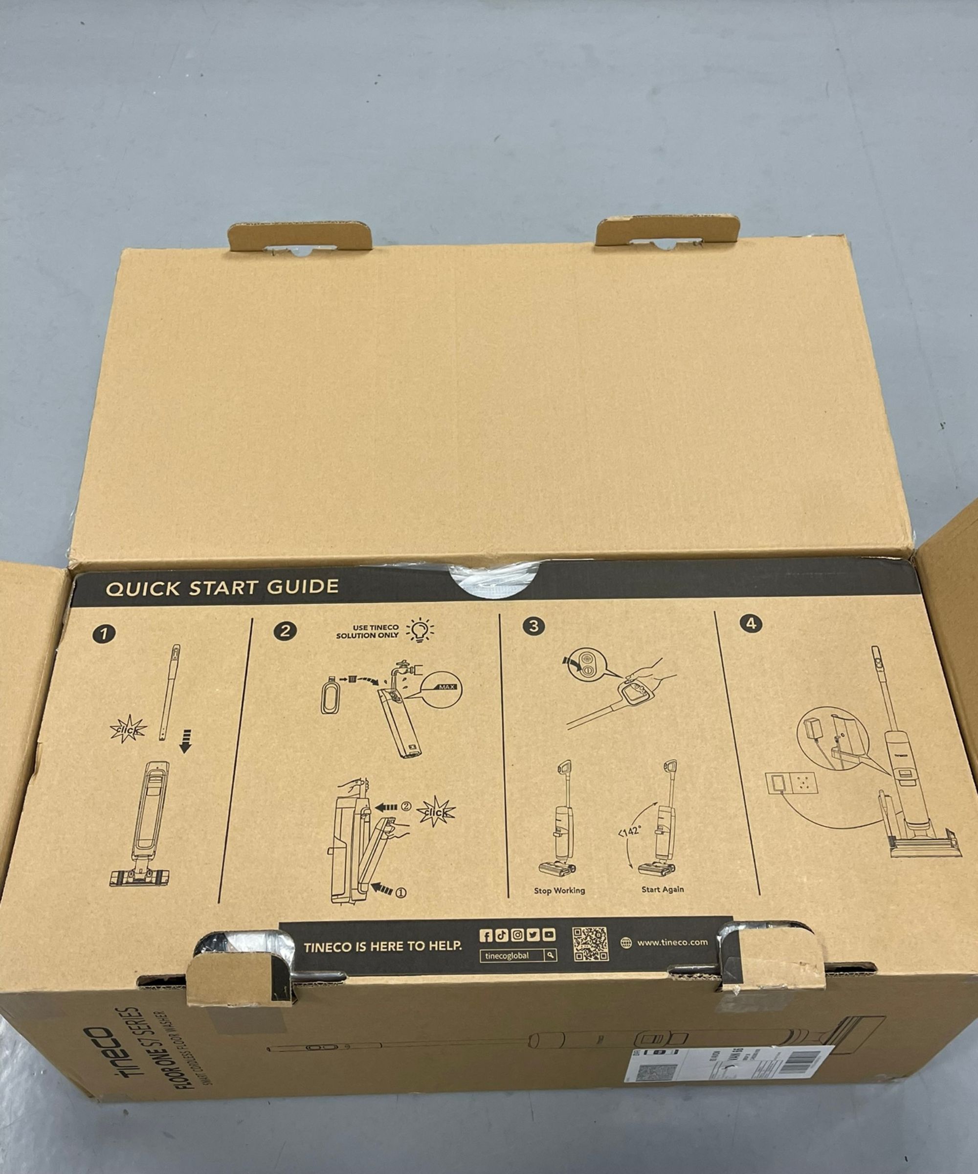 Tineco Floor One S7 Pro Cordlessless Wet Wet Vacuum's Cardboard Box, partiellement ouvert montrant un guide de démarrage rapide sur la façon d'utiliser l'aspirateur, sur le plancher gris dans le futur centre de test