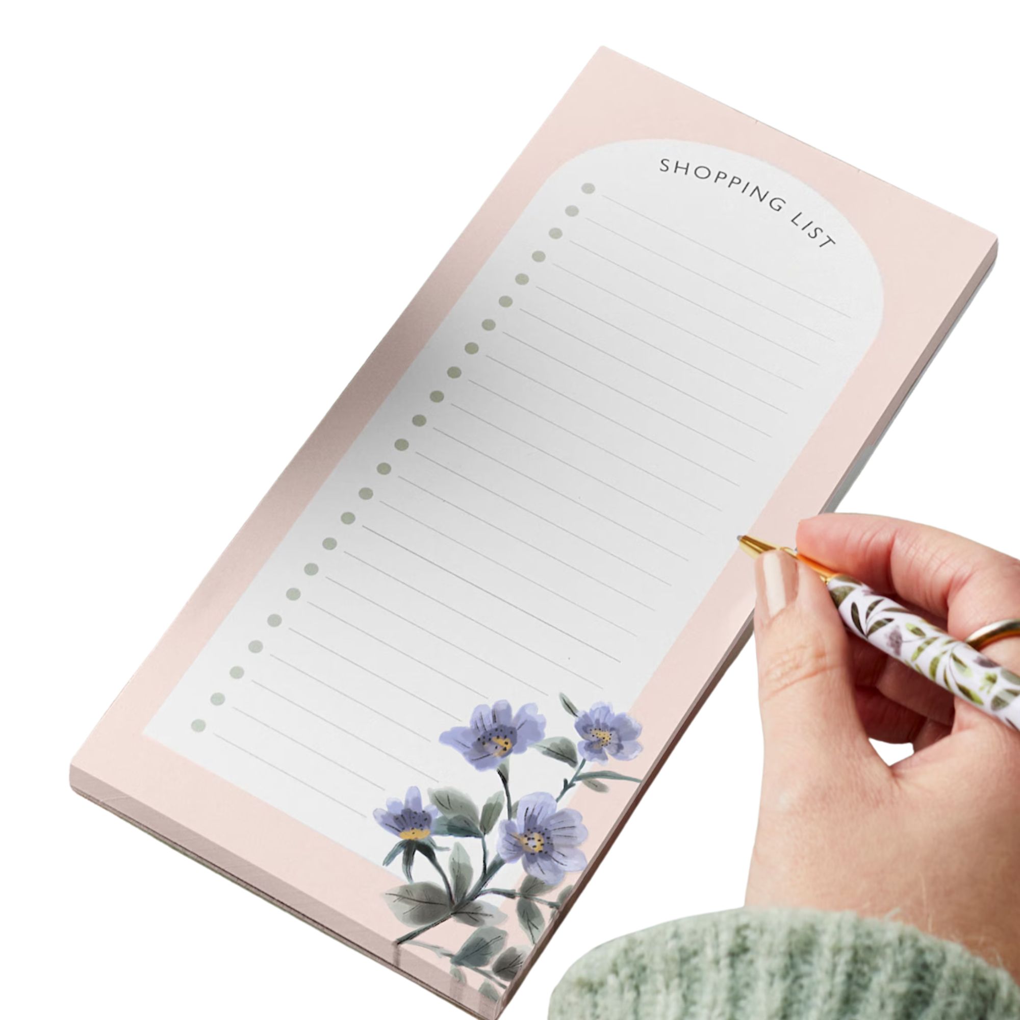 Une main avec une manche à sauteur en laine verte sage tenant un stylo floral blanc et or sur une liste de courses rose pâle et blanc avec des détails de fleurs violettes dans le coin inférieur droit