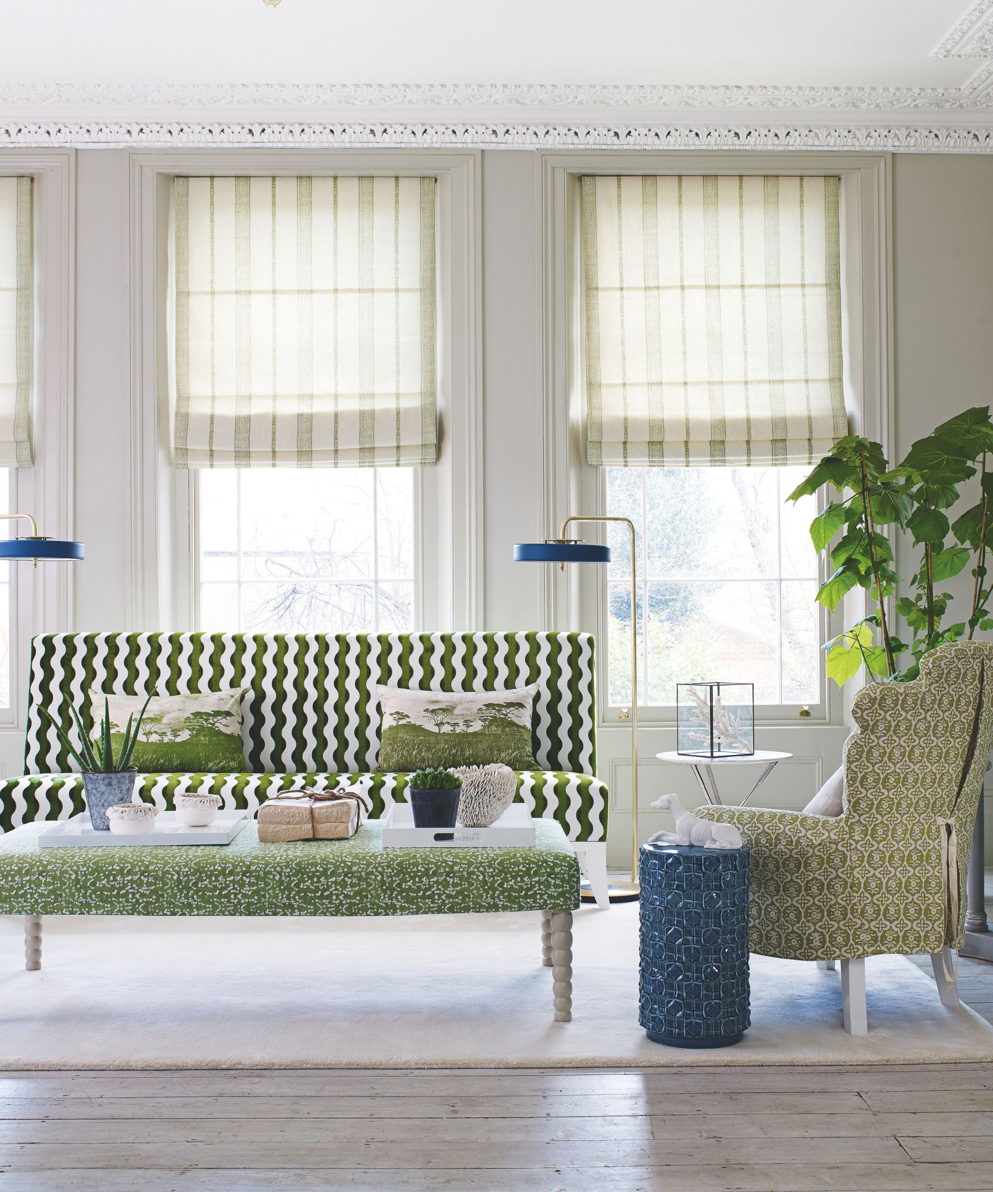 Un salon lumineux avec de grandes fenêtres avec des stores géométriques et un canapé géométrique à motifs, un fauteuil et un repose-pieds rembourrés en motifs verts, deux lampes en or et bleues autonomes, une table basse cylindrique bleue et une grande plante