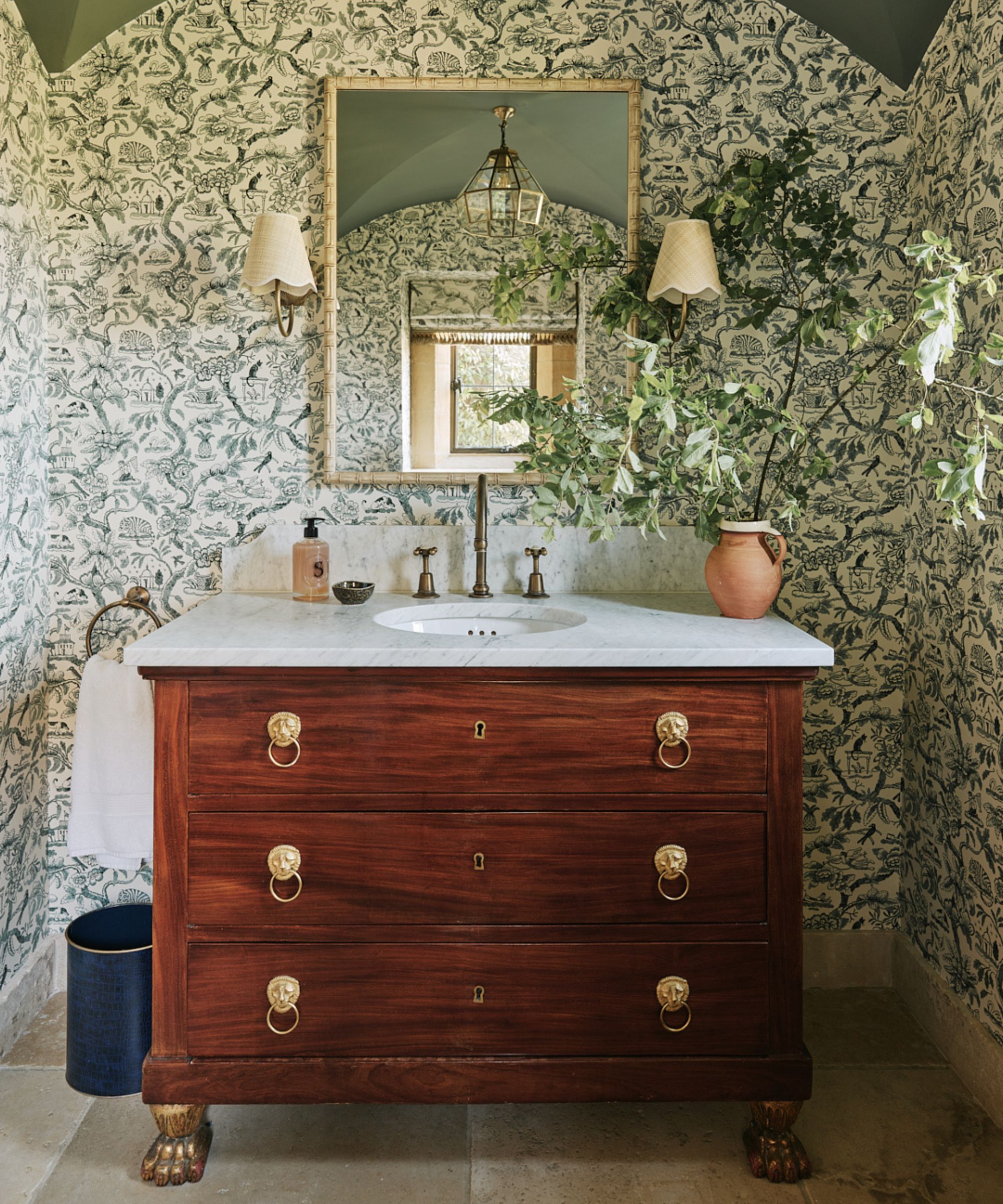 Petite salle de bain peint avec du papier floral vert et blanc avec un évier de vanité vintage qui a un petit vase en terre cuite rempli de branches