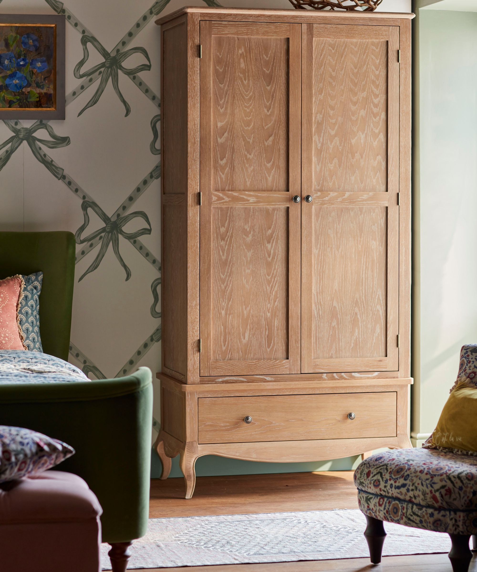 Closet à deux portes en pin autoportant avec quincaillerie en laiton, lit en velours vert, tapis géométrique, ottoman rose blush au pied du lit et fauteuil coloré et oreillers. Le fond d'écran est crème avec des gros arcs verts sages