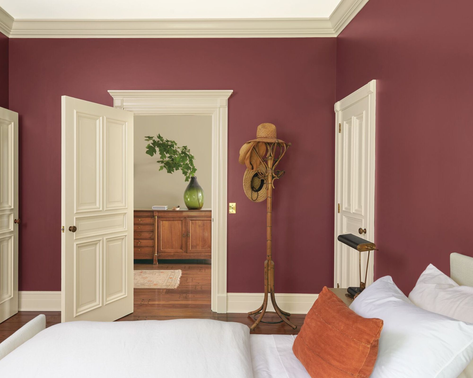 Une chambre avec un lit avec des draps blancs et un coussin orange, un manteau dans le coin avec des chapeaux dessus, des murs peints de couleur rouge baie et les plinthes et les portes sont peintes en blanc cassé.