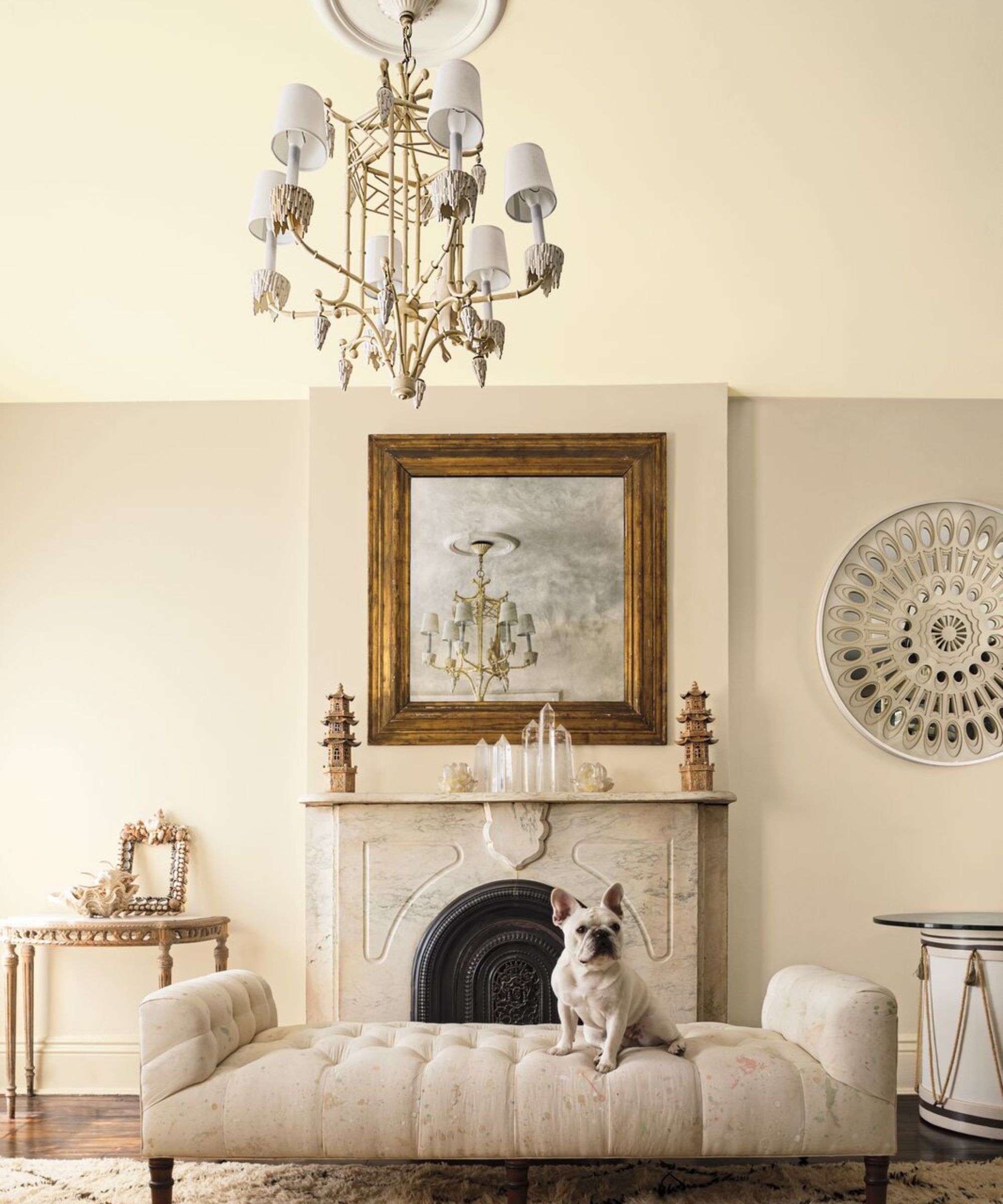 Un salon avec des murs peints dans une couleur neutre chaude. Il y a une cheminée en marbre avec un miroir encadré doré suspendu au-dessus. Un banc neutre rembourré est devant le feu avec un bouledogue français assis sur le dessus.