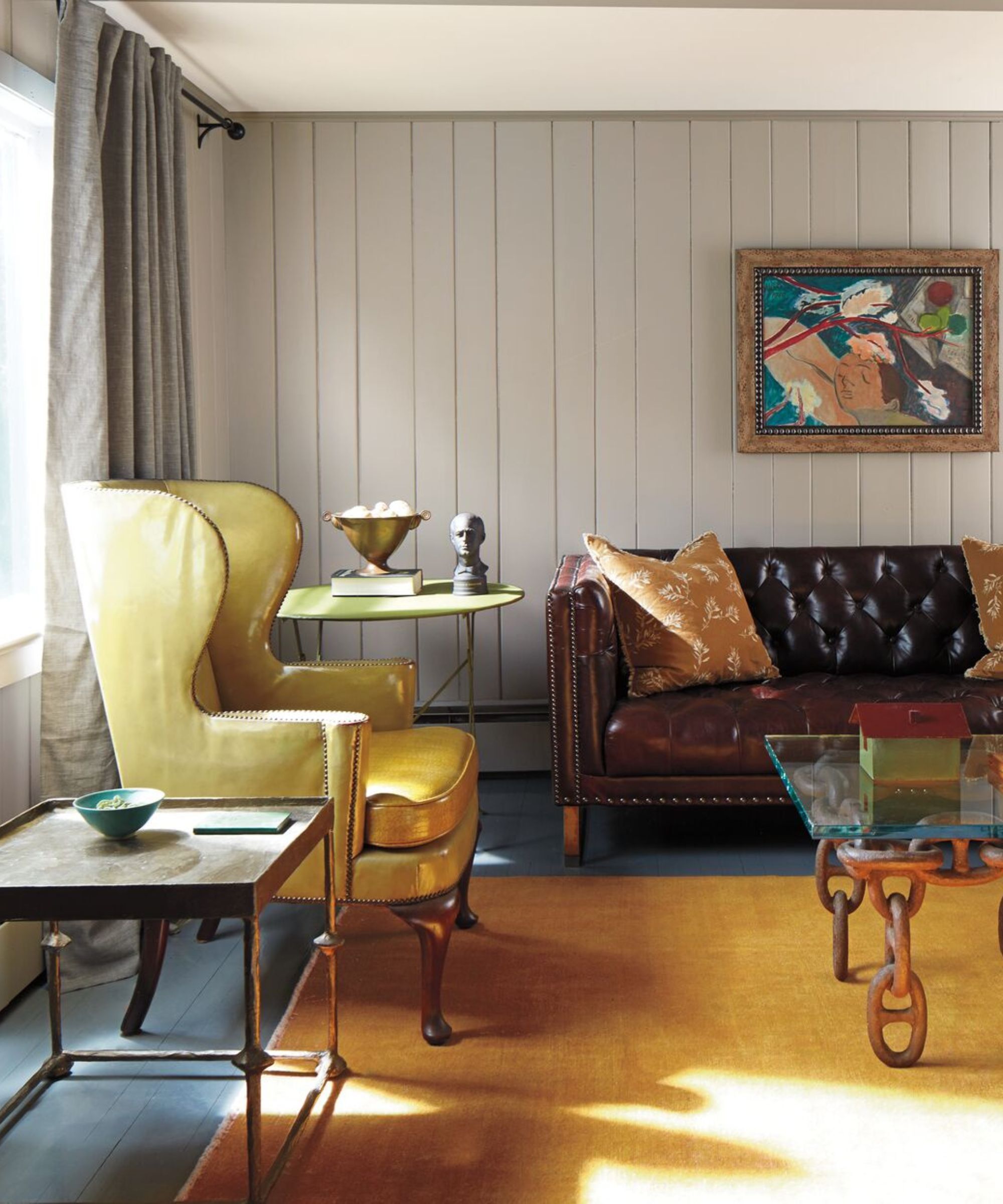 Un salon avec un mur lambrissé peint dans un gris neutre. Il y a une peinture colorée suspendue au mur et un canapé en cuir et un fauteuil en cuir. Il y a un tapis de couleur moutarde sur le sol et une table basse de style industriel.