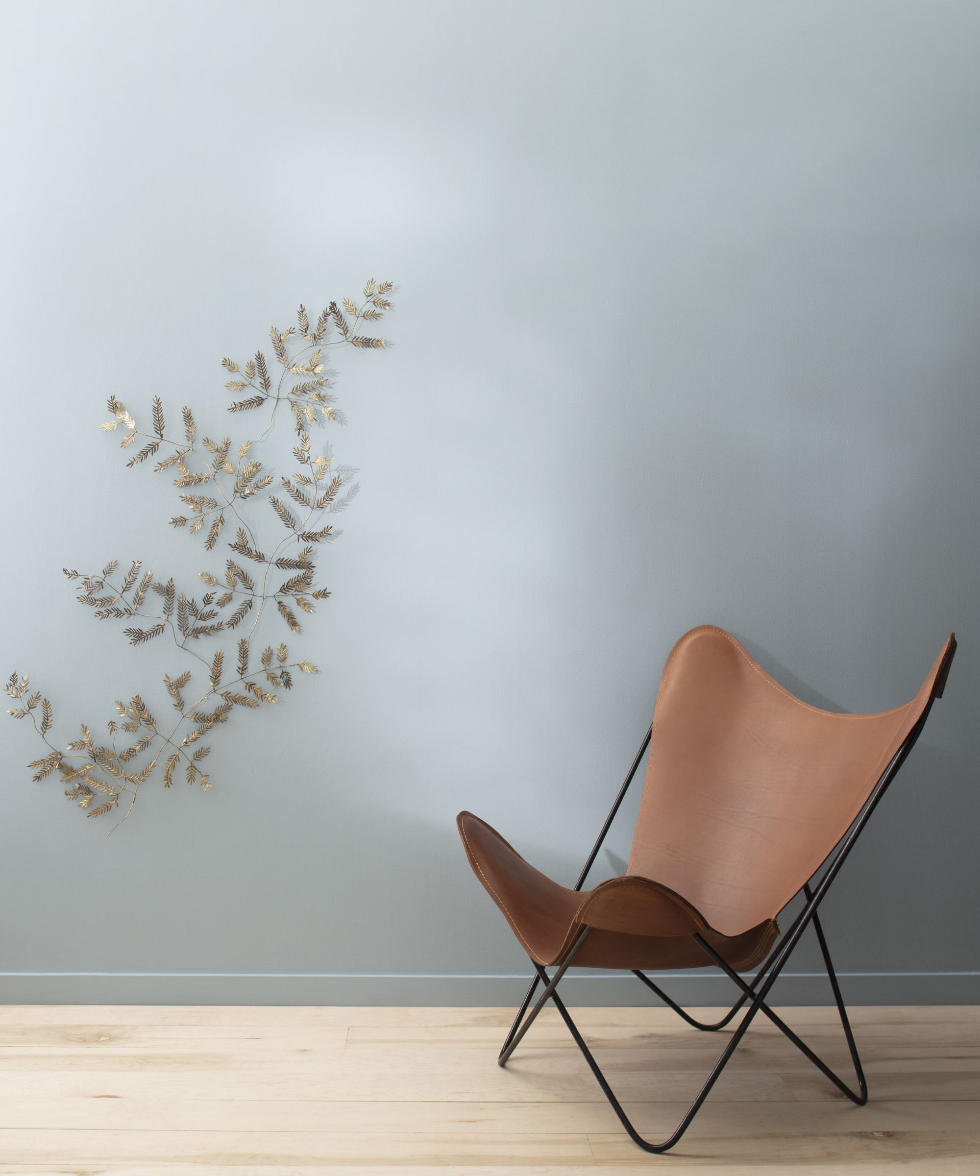 Un mur peint dans une peinture grise froide, avec un mur décoratif suspendu rappelant une branche avec des feuilles. Une chaise en cuir est devant le mur.