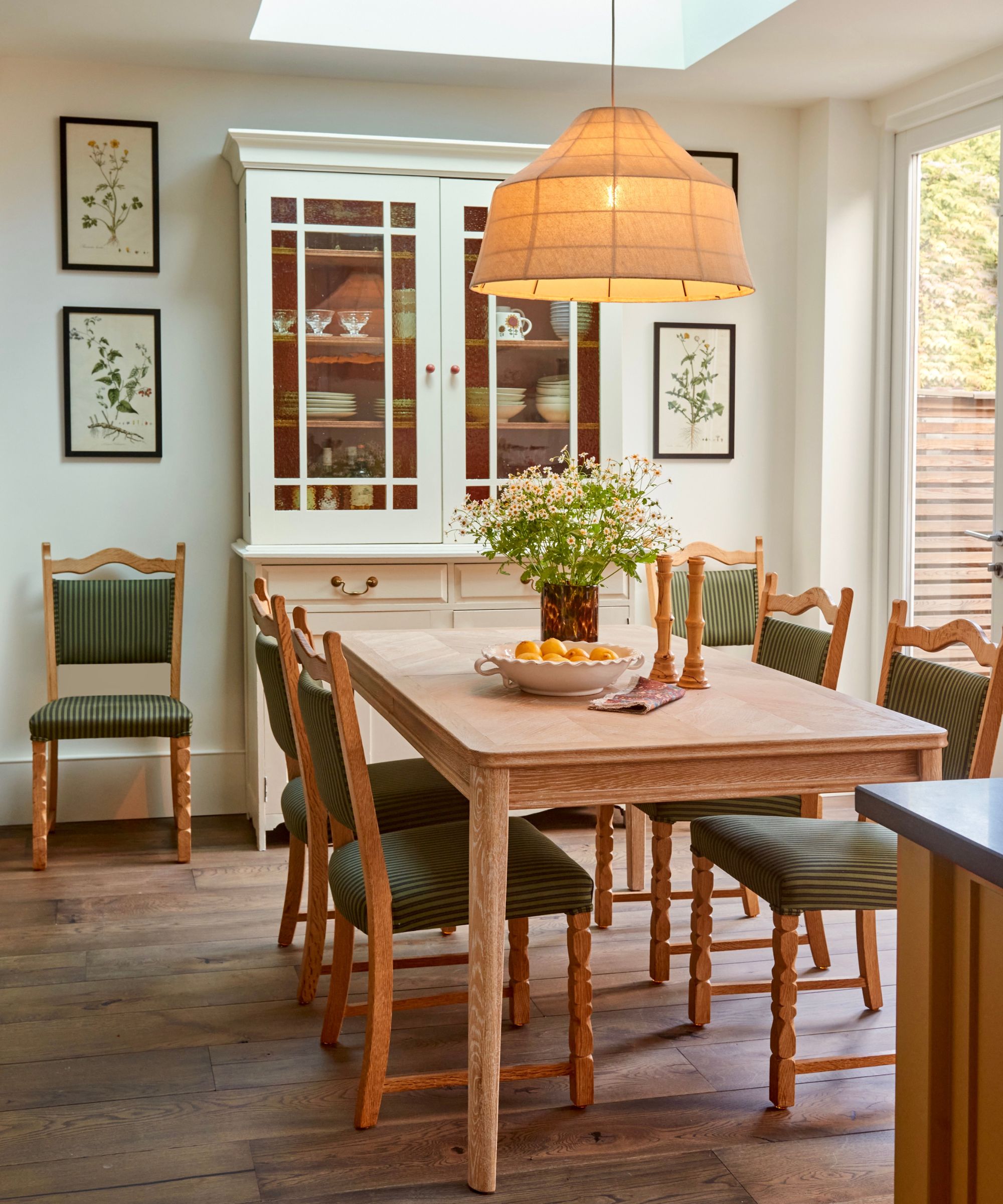 Salle à manger avec table et chaises en bois vintage, pendant, armoire en bois avec assiettes et verres et imprimés botaniques vintage