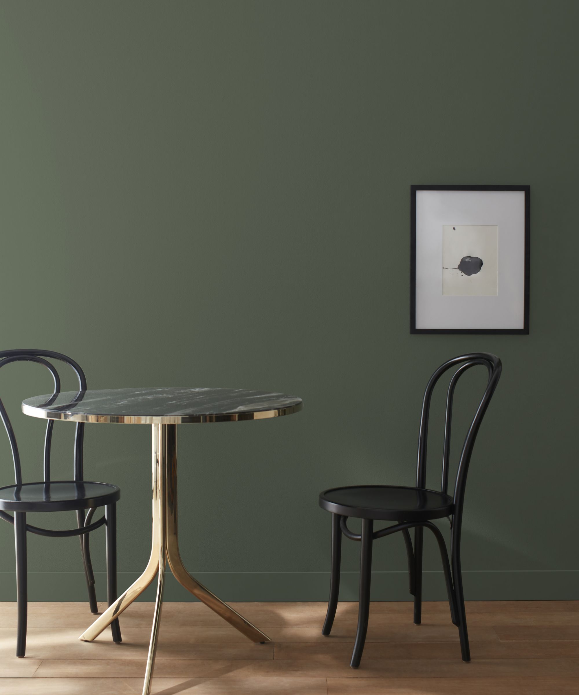 mur vert foncé avec table circulaire et deux chaises noires, image noire encadrée sur le mur