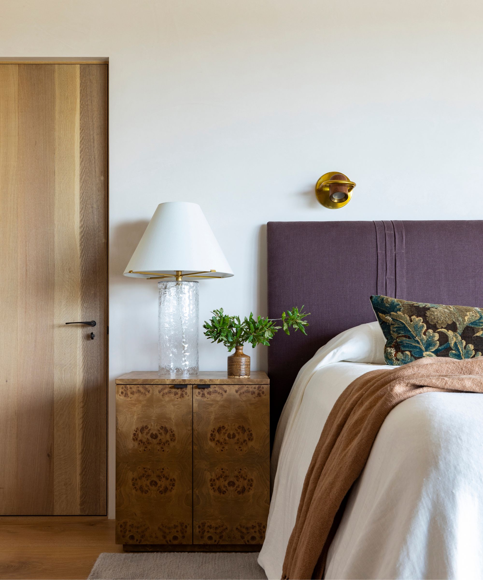 Chambre neutre avec une tête de lit violette en lin, literie en lin blanc, un oreiller de tapisserie et une table de chevet en bois de Burl de style vintage