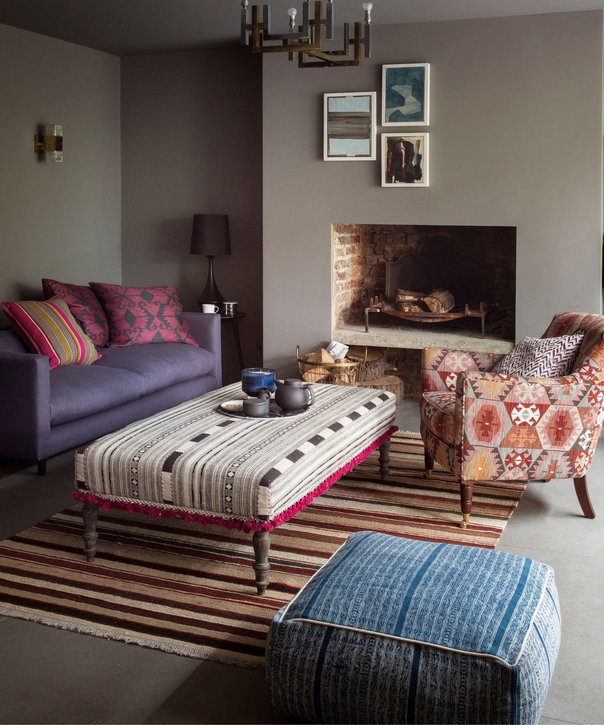 Un salon brun neutre avec un pouf à motifs contrastés, une chaise et du pouffe, un tapis brun rayé et un canapé violet avec des coussins roses vifs et trois images encadrées en cadres blancs sous un pendentif industriel métallique