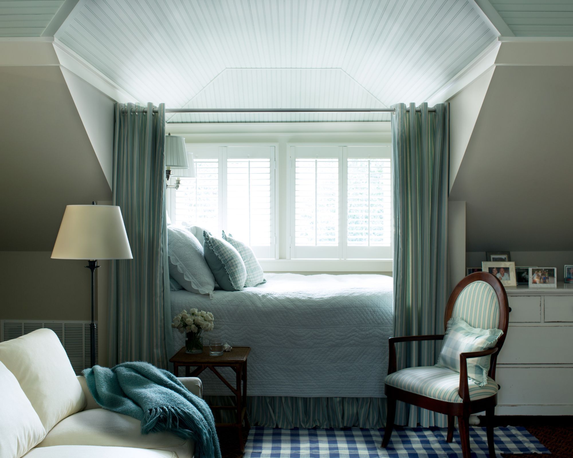 Une chambre bleu pâle avec un canapé à crème, une chaise à rayures bleues et un lampadaire. Il y a un tapis à carreaux bleus sur le sol et un lit avec un linge de lit blanc et bleu traditionnel. Il y a des rideaux bleu pâle entourant le lit. Les murs sont peints dans de la peinture grise.
