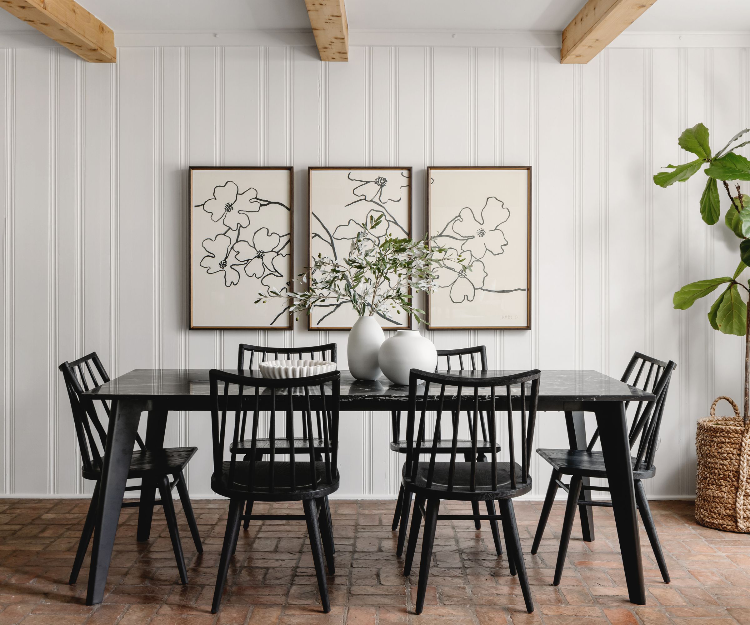 Salle à manger avec murs en bois blanc, trio d'art mural, table et chaises en bois noir et feuillage en vase