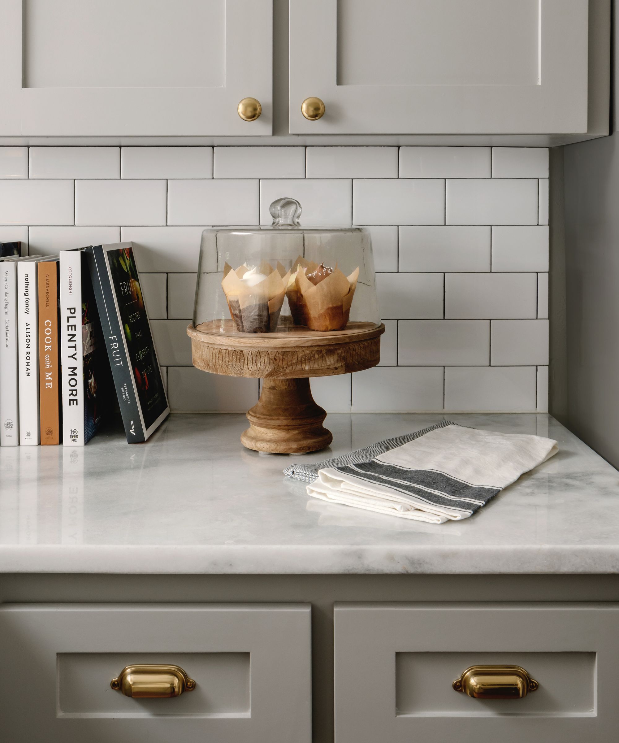 Une cuisine cottage avec des armoires grises, des comptoirs en marbre, du matériel en laiton, un support à gâteau en bois et une pile de livres de recettes