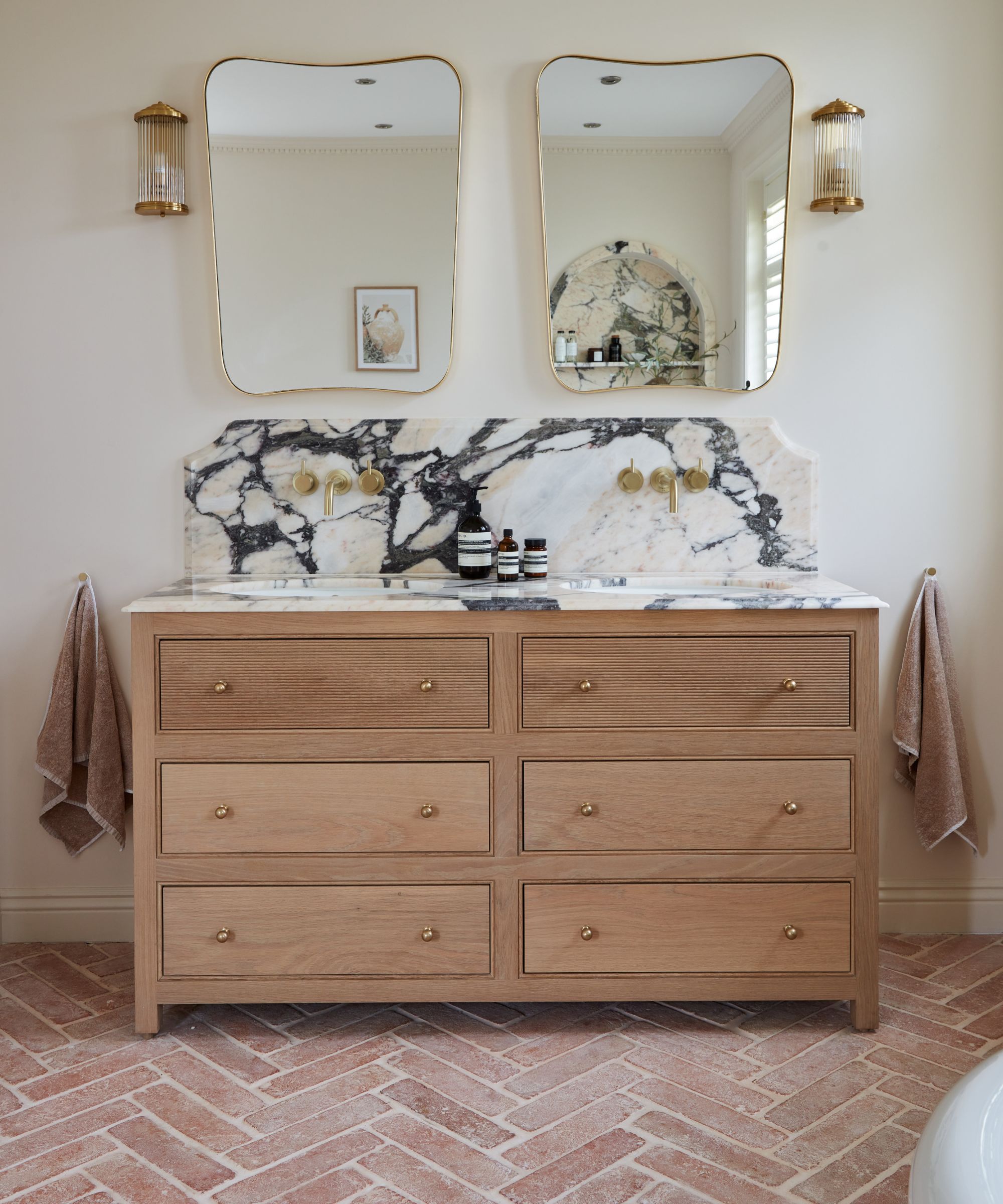 Une salle de bain blanche avec des carreaux de sol en terre cuite et une vanité en bois avec un comptoir en marbre et des miroirs de vanité en or