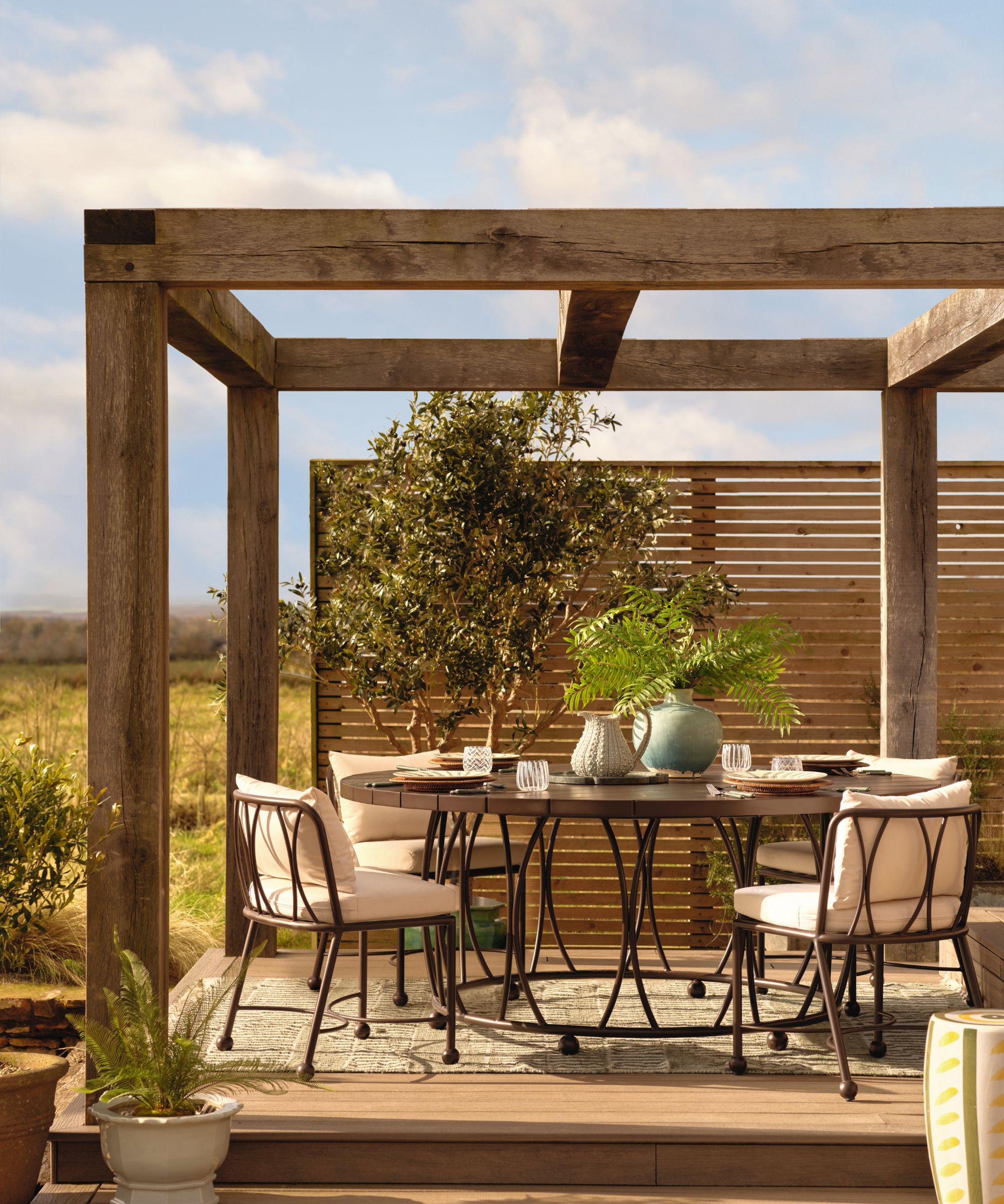 Une arrière-cour avec une pergola en bois, un mobilier d'extérieur en métal avec une vaisselle dessus et un tapis extérieur sur le sol