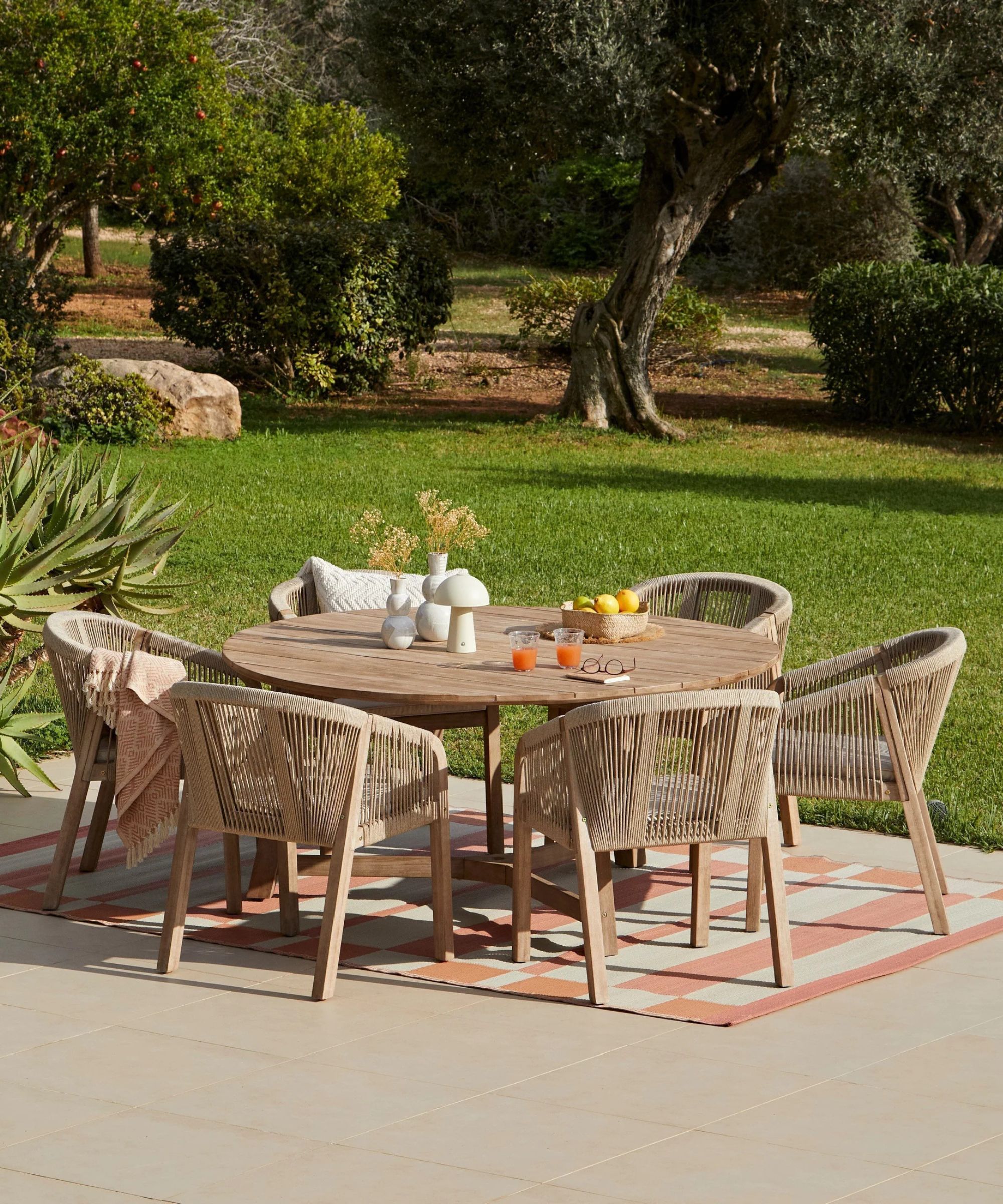 Une zone d'arrière-cour avec un ensemble de meubles d'extérieur en bois, un tapis extérieur rose et blanc et une pelouse et des arbres