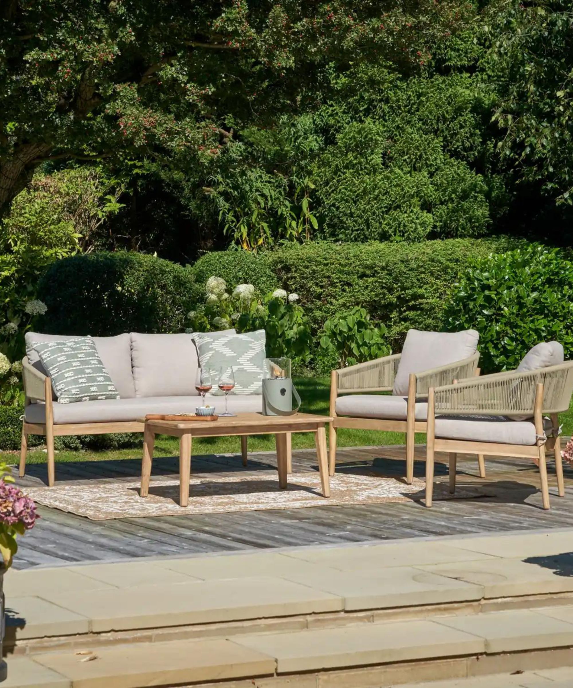 Une cour à l'arrière avec un ensemble de meubles d'extérieur gris et en bois, un tapis extérieur beige et des arbustes verts autour