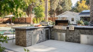 Outdoor kitchen in stone with stainless steel cabinets and grill, paved patio and swimming pool and buildings beyond