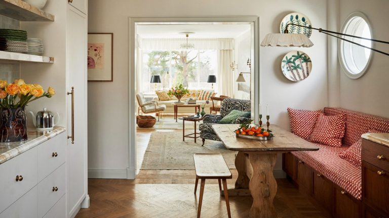 An archway from a kitchen-diner with a red banquette bench and wooden table, into a brightly lit living room.