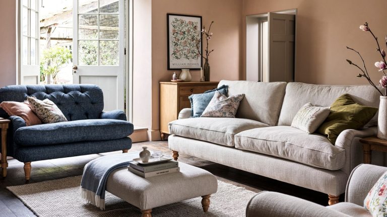 A pink living room with a cream sofa and blue sofa, around a small cream footstool coffee table.