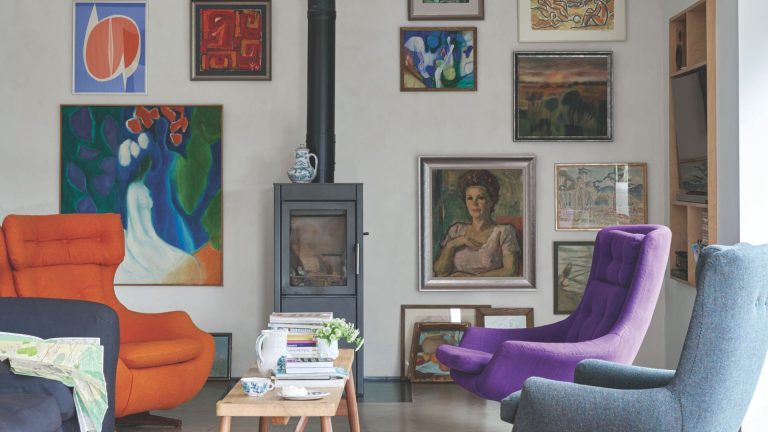 A gallery wall painted white with lots of artwork and a wood burning stove. There are colorful modern chairs and a wooden coffee table.