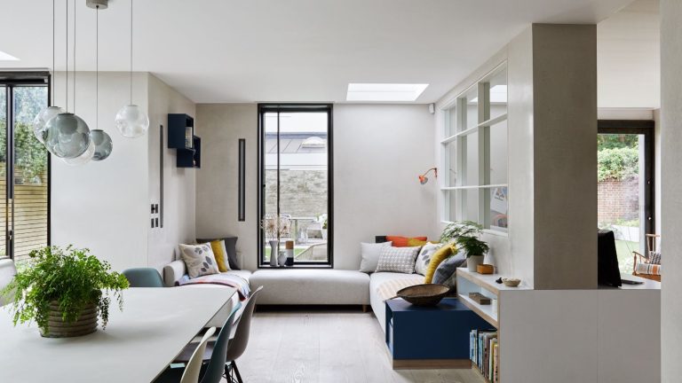 A bright seating area filled with natural light from large windows, with light wooden flooring, a cream corner sofa, and a cream table with glass pendant lights hanging above