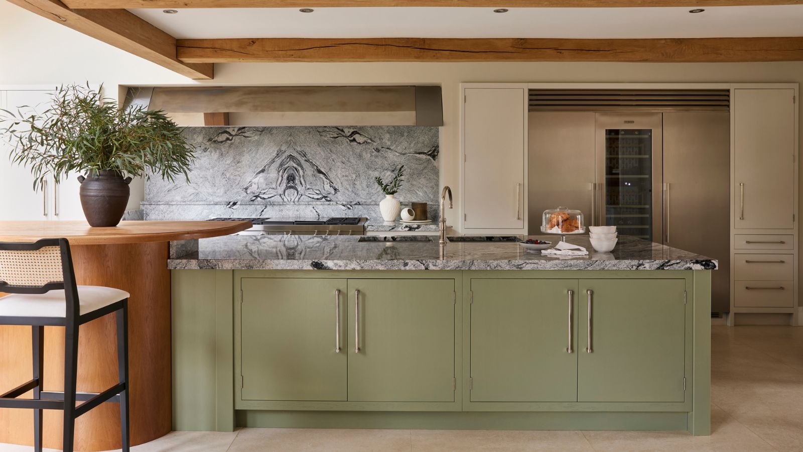 Une cuisine avec un grand îlot de cuisine peint en vert et un comptoir en marbre, intégrée dans une grande table de cuisine orange avec des tabourets de bar noirs. Un réfrigérateur-congélateur du sol au plafond intégré à l'arrière-plan. 