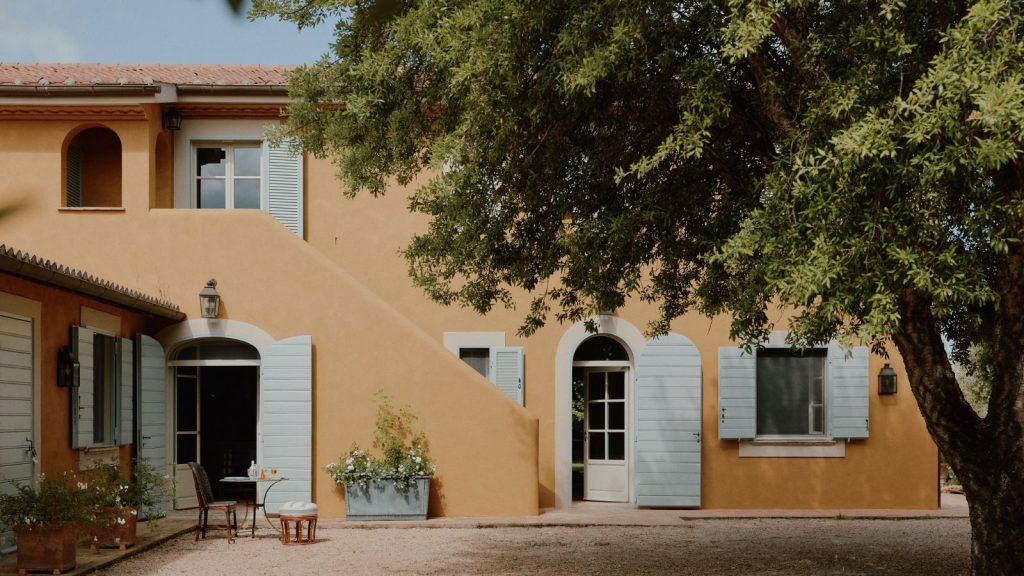 Comment un créateur britannique a rassemblé les différents goûts d’un couple qui voulait créer la maison de rêve à l’épreuve du futur dans la campagne idyllique italienne Exterior of a pale orange house with pale blue shutters and a large tree in front
