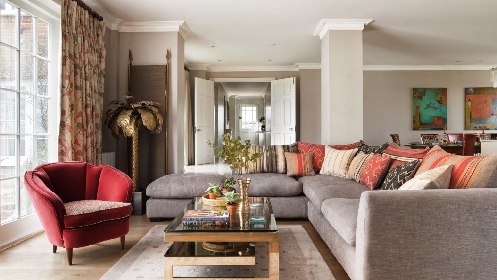 Large living room with open double doors revealing entryway. Coffee table with ornaments, L-shaped gray sofa and red armchair in foreground, with abstract paintings and pillar in the living room.