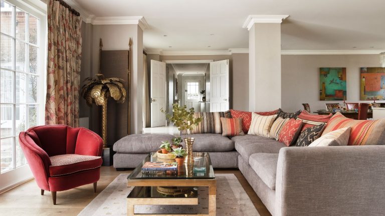 Large living room with open double doors revealing entryway. Coffee table with ornaments, L-shaped gray sofa and red armchair in foreground, with abstract paintings and pillar in the living room.