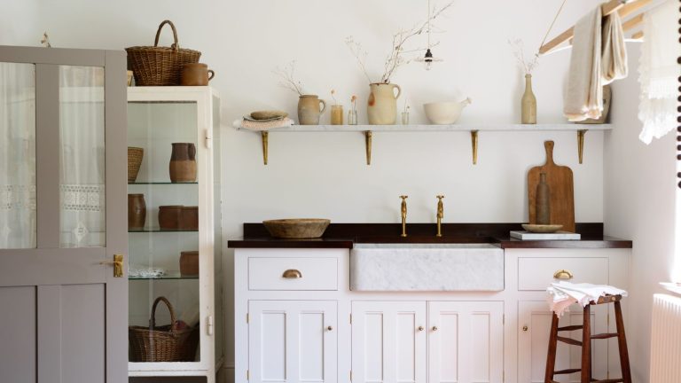 Comment faciliter le linge pliant – 5 astuces du timing aux tactiques pour vous aider à travailler plus intelligemment, pas plus difficile White cottage style laundry room with shaker cabinets, marble sink, brass fixtures, glass cabinet with woven baskets and natural material storage pots. There is a shelf above the sink, and a line drying apparatus mounted to the ceiling on the right