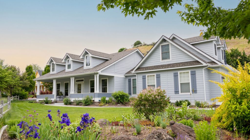 A light gray house with a blue sunset sky above it and a green lawn in front of it with purple flowers