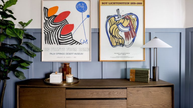 Entryway with blue wood panelled wall and wood console with lamp and books and two vintage posters
