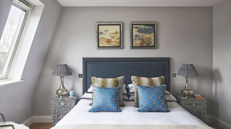 A gray bedroom with a double bed with blue headboard, white bedsheets and blue, white and green patterned cushions. On either side of the bed are black and white patterned bedside tables, and above are two framed artworks in silver framed. To the left is a slanted window.