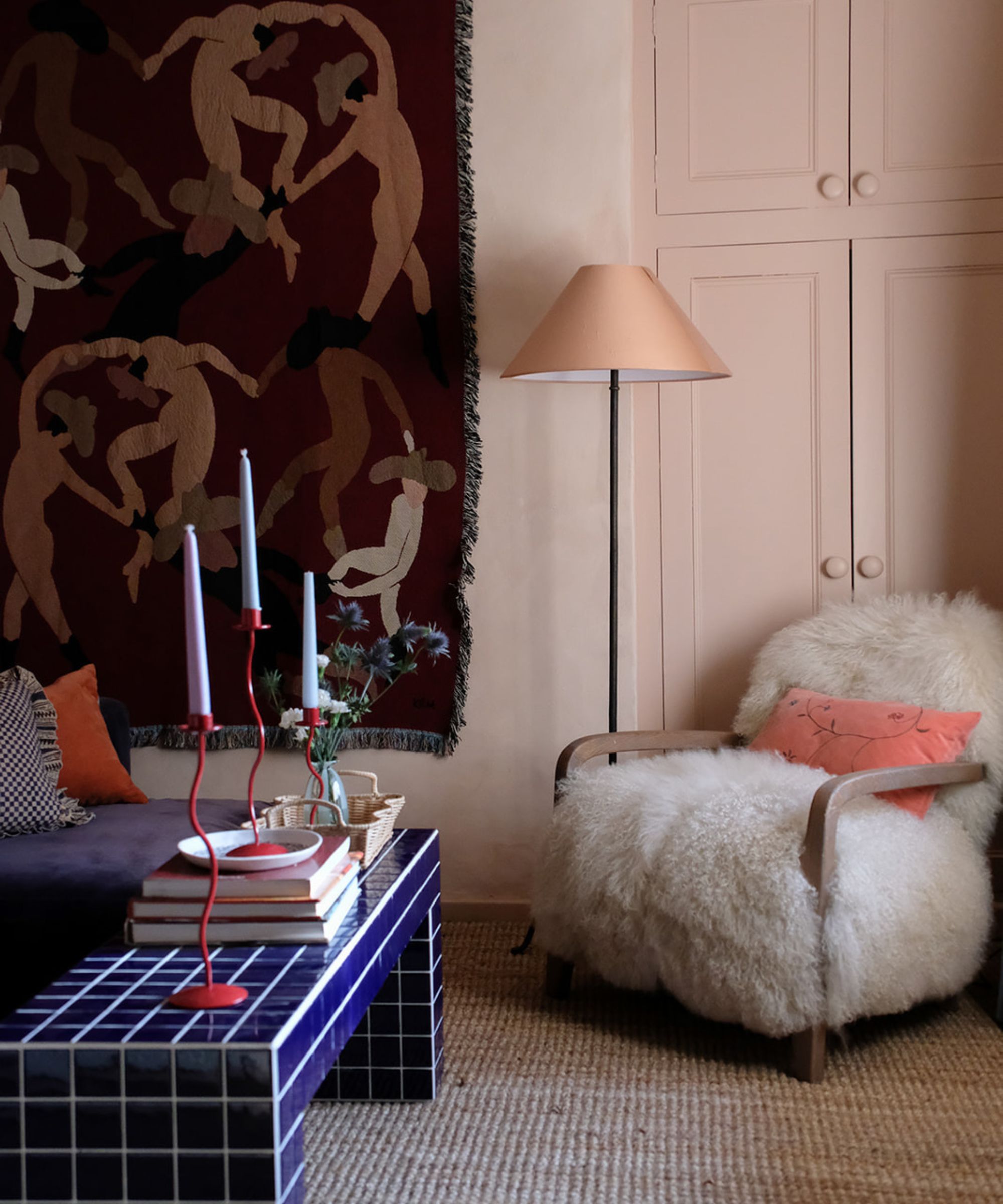Salon éclectique avec armoire rose, fauteuil moelleux et table basse carrelée bleue et tapisserie suspendue
